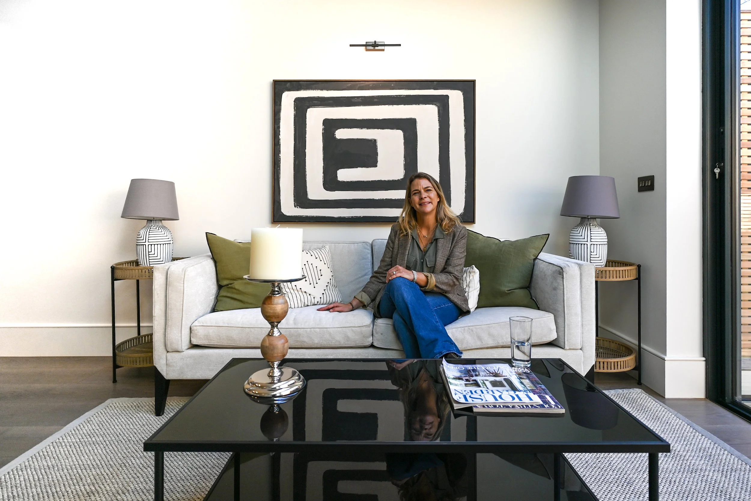 Woman sitting on a light-colored sofa in a modern living room, with two side tables, lamps, and a black-and-white abstract painting behind her.