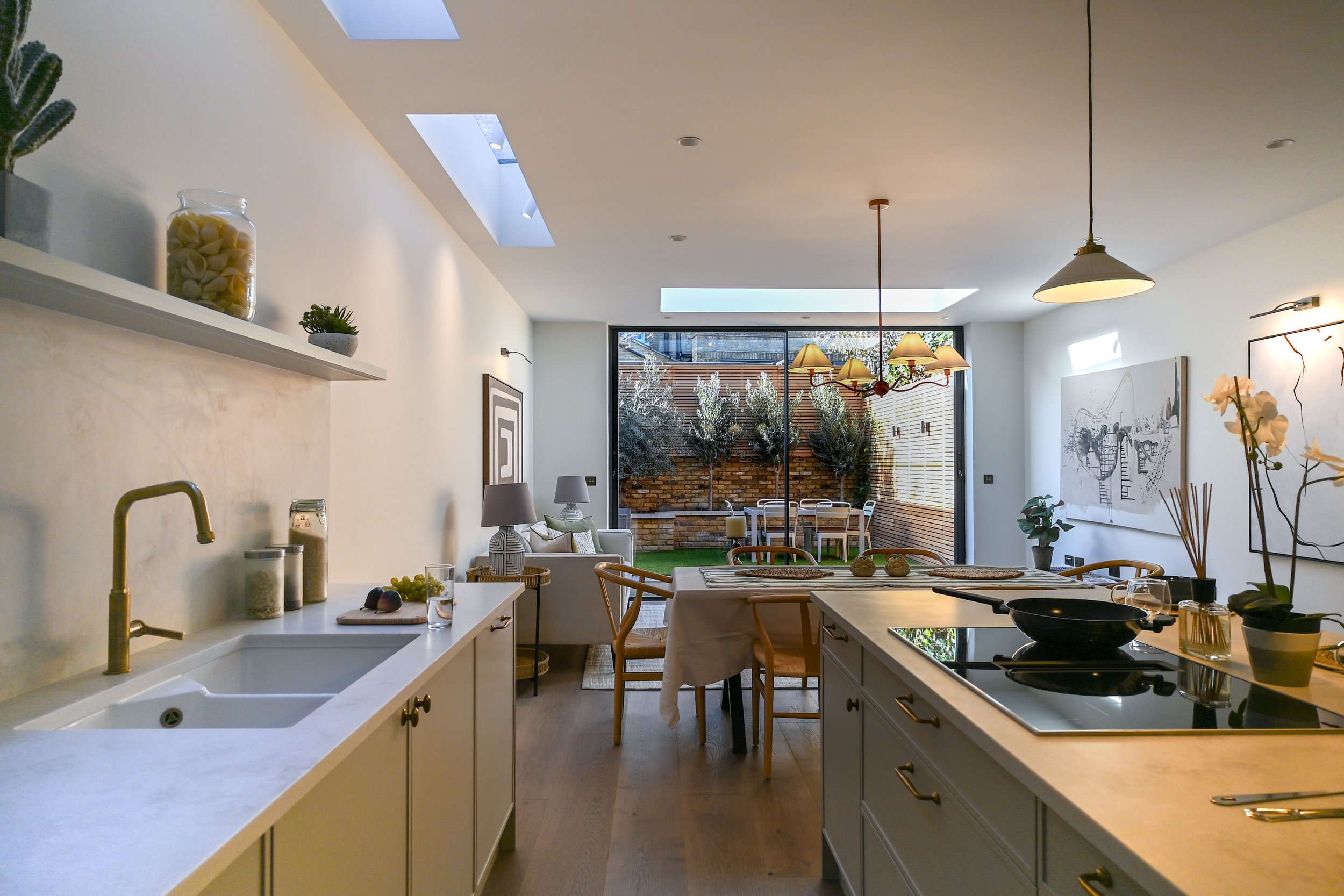 Open-plan kitchen and dining area with large glass doors leading to a garden patio, featuring a kitchen island with a stovetop, a dining table with chairs, and a sitting area with sofas and lamps.