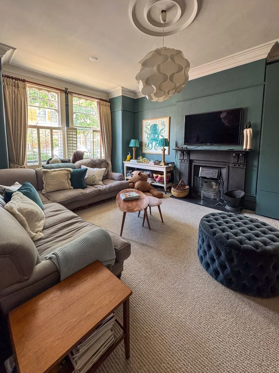 A cozy living room with beige sofas, decorative pillows, a small wooden coffee table, a blue velvet tufted ottoman, and a fireplace with a mounted television. Large windows with curtains allow natural light to fill the space, complemented by tree vie