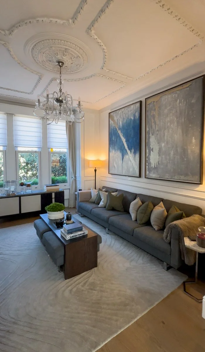 Living room with a large gray sofa, abstract art on the wall, a chandelier, large windows with white blinds, and a modern coffee table.