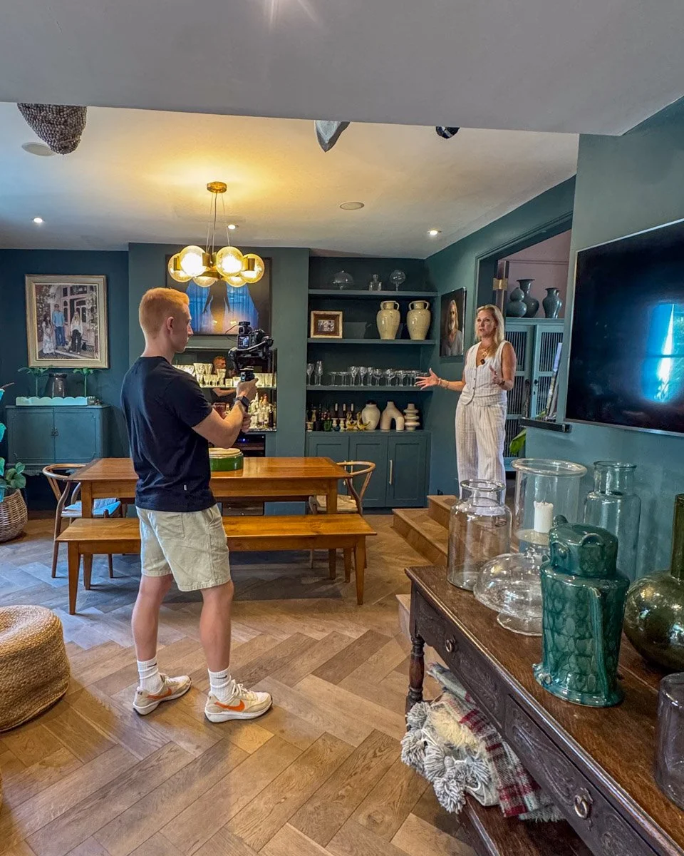 A young man records a woman giving a presentation in a living room with shelves of ceramics and books, a wooden dining table, and decorative items.