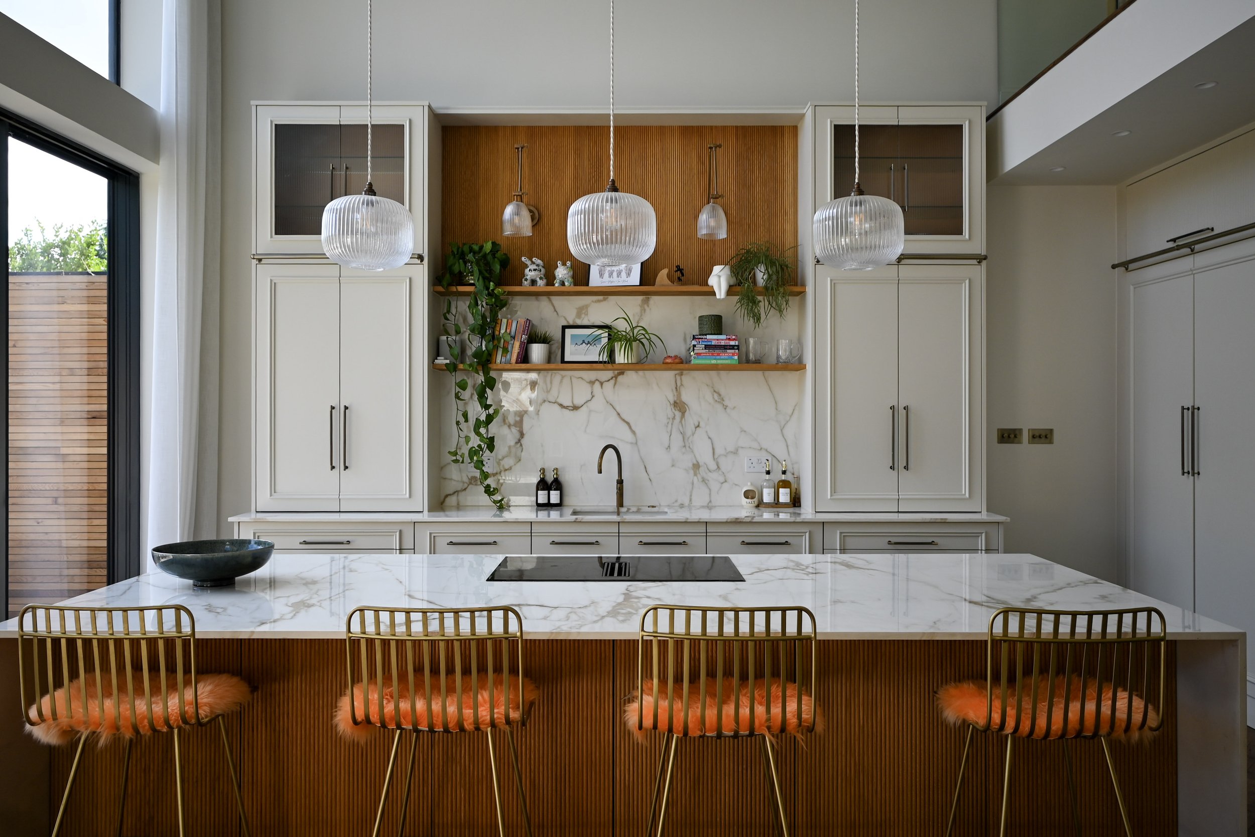 Modern kitchen with white cabinetry, marble countertops, and pendant lighting. An island with a marble top and four gold chairs with orange furry cushions. Open shelves with plants, books, and decorative items. Large sliding glass door to the left.