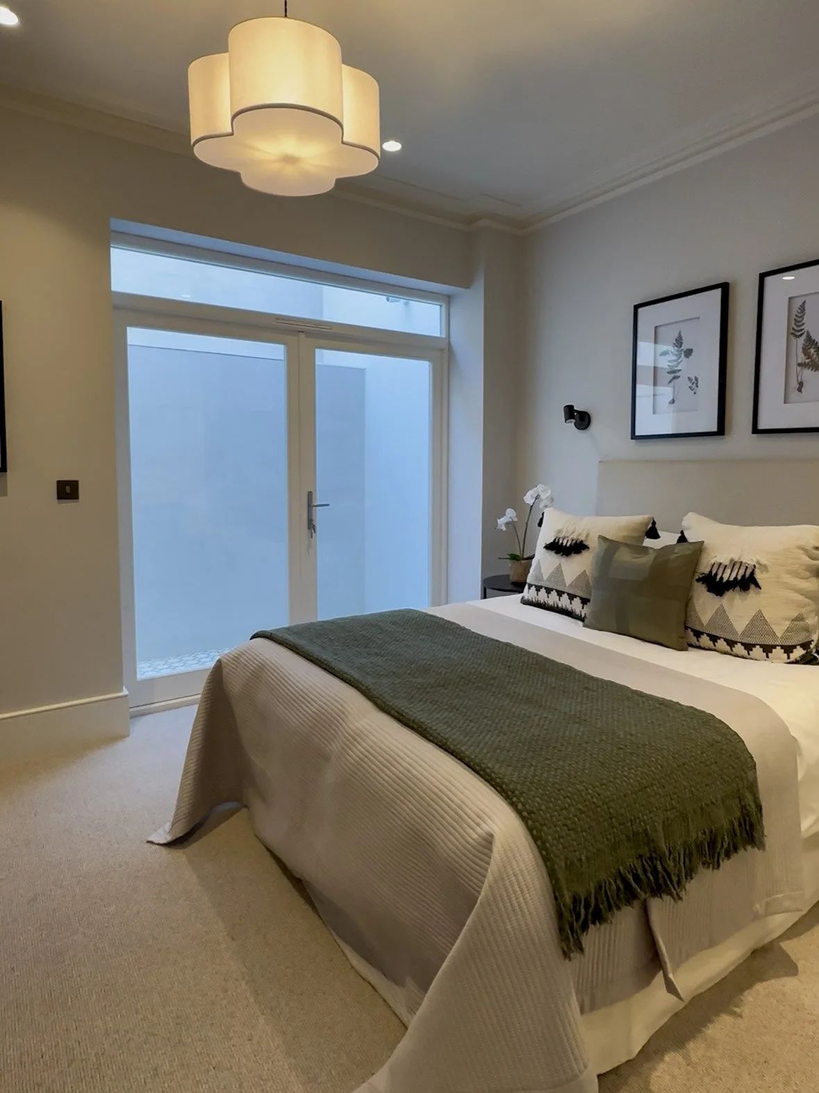 A bedroom with a bed covered in neutral-toned bedding and pillows, a sliding glass door, framed botanical prints on the wall, a black wall-mounted reading light, and a ceiling light fixture.