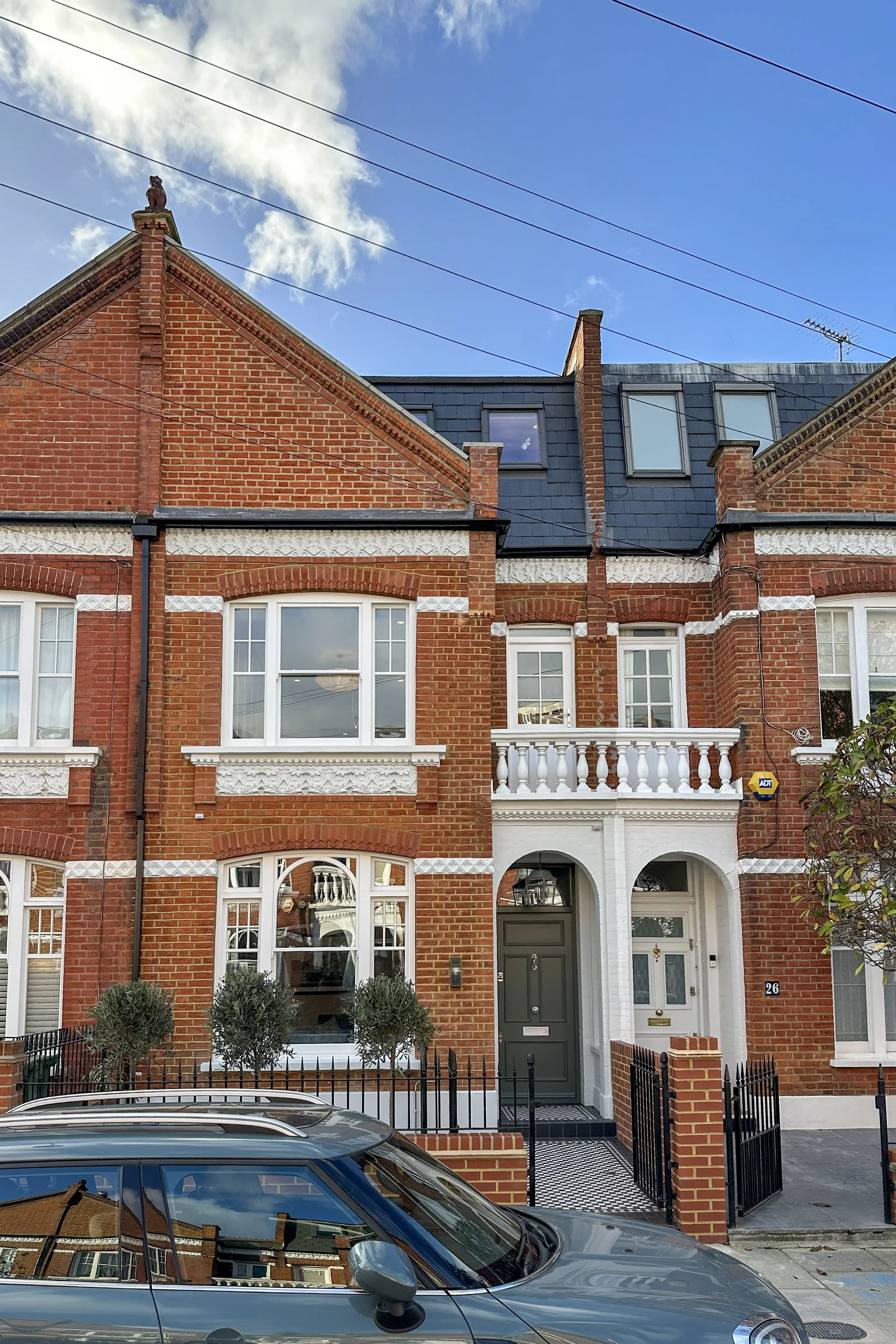 A row of brick townhouses with decorative white accents, front gardens, and a parked car in the foreground under a partly cloudy sky.