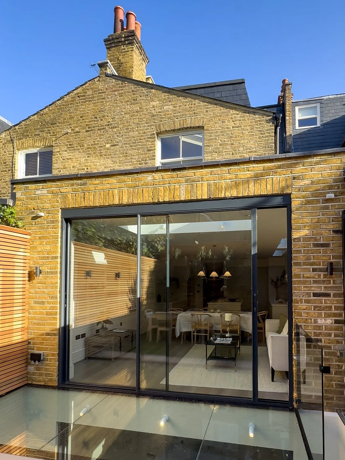View of a house with a brick exterior, large sliding glass doors, and a small enclosed patio area with a glass railing. The interior is a modern living and dining space visible through the doors.