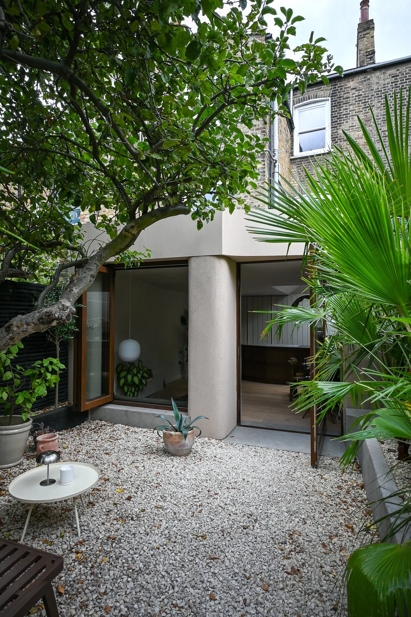 A small urban backyard garden with white gravel ground, potted plants, and a modern house with large glass doors. There are trees and large leafy plants surrounding the space.