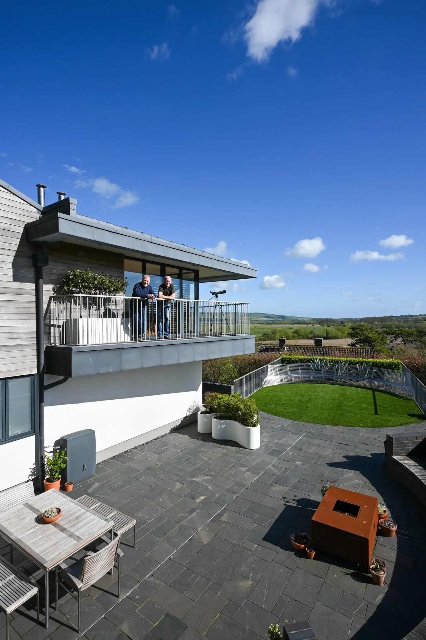 A spacious outdoor patio with gray tile flooring, a white modern building with large windows and balcony, potted plants, and a green lawn with a hilly landscape in the background under a bright blue sky with clouds.