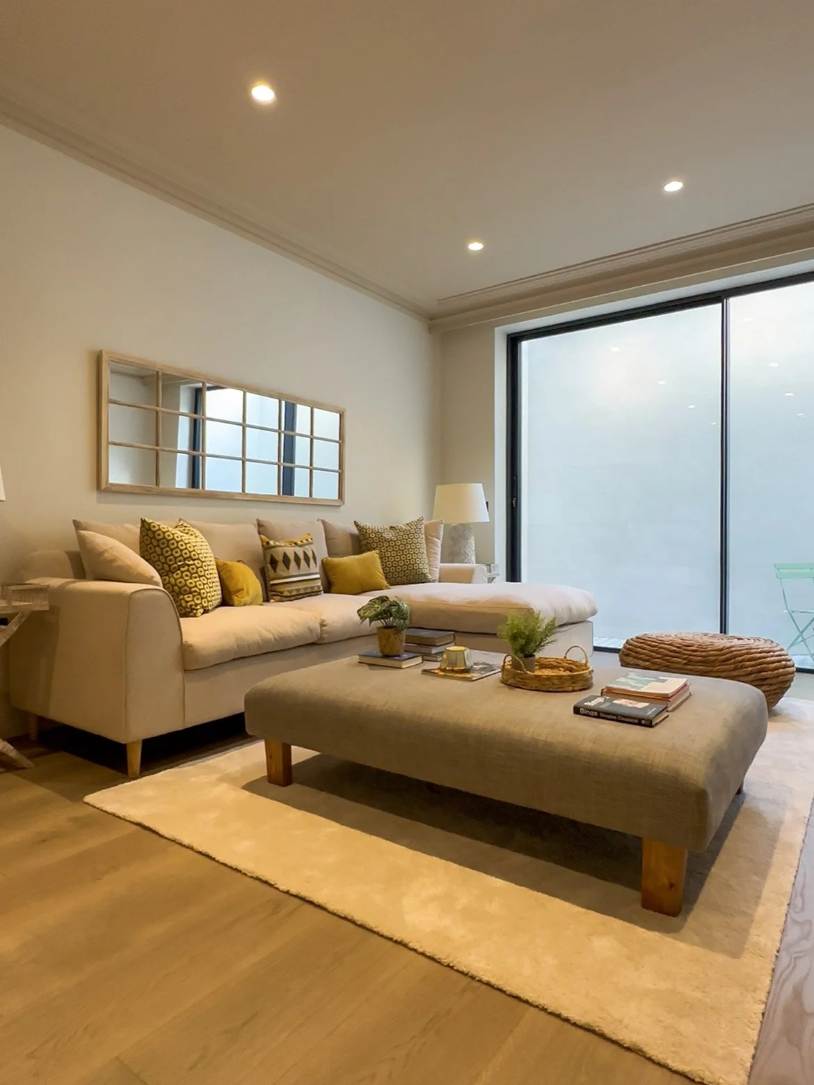 Living room with beige sofa, six patterned throw pillows, a beige ottoman with books, plants, and a tray, a cream textured pouf, and a large window with frosted glass. The room has recessed ceiling lights, a white lamp, a framed mirror, and a cream area rug on wooden flooring.