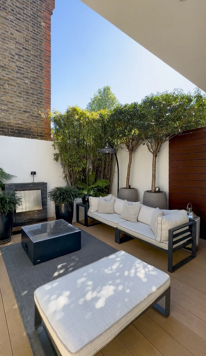 Outdoor patio with a white sofa, potted trees, plants, a black coffee table, and a cushioned ottoman, with wooden flooring and a white wall background.