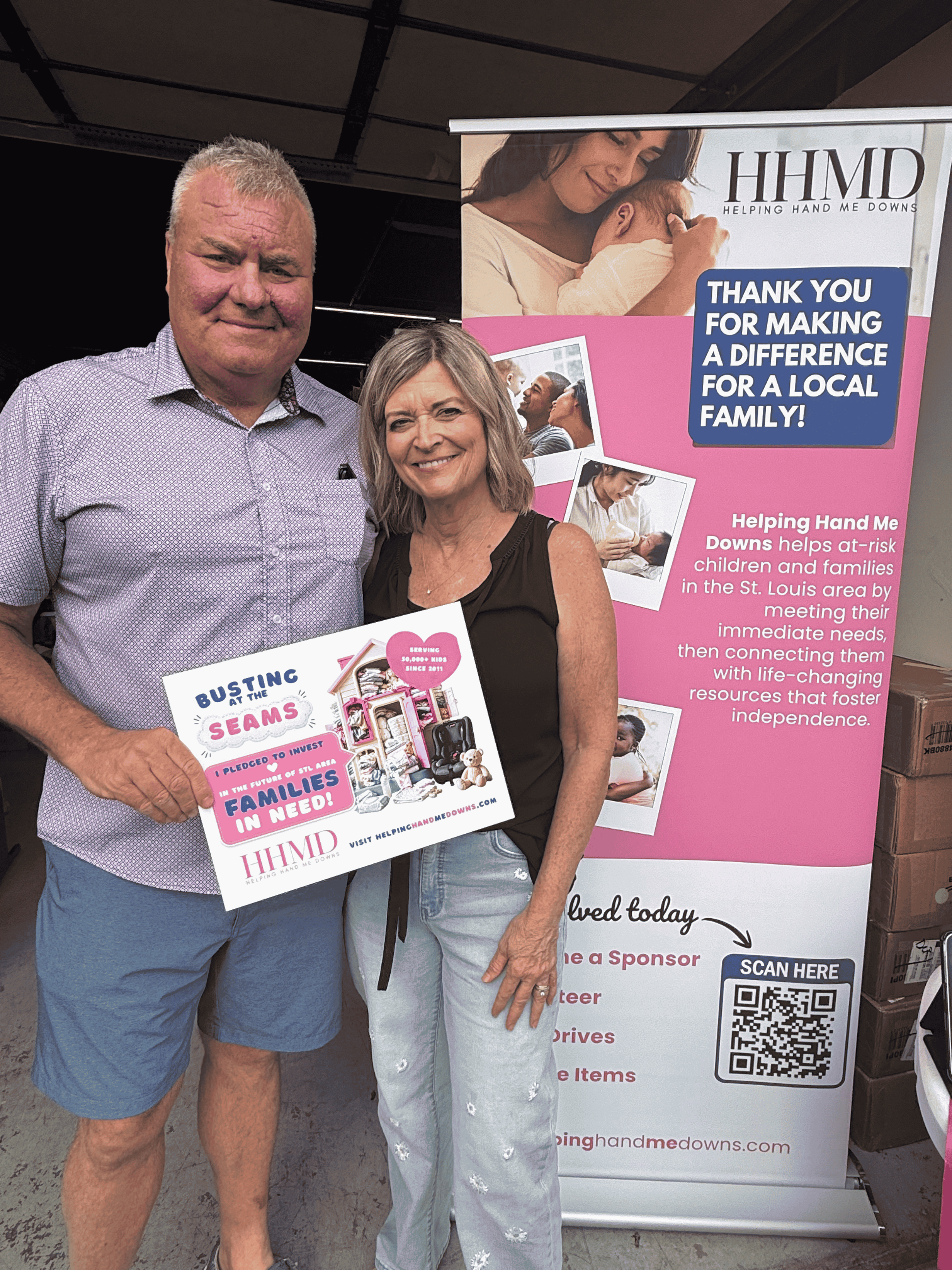 A smiling man and woman standing together in front of a pink and white informational display about Helping Hand Me Downs, holding a donation request sign. The display features images of children and families. Helping Hand Me Downs Building Campaign