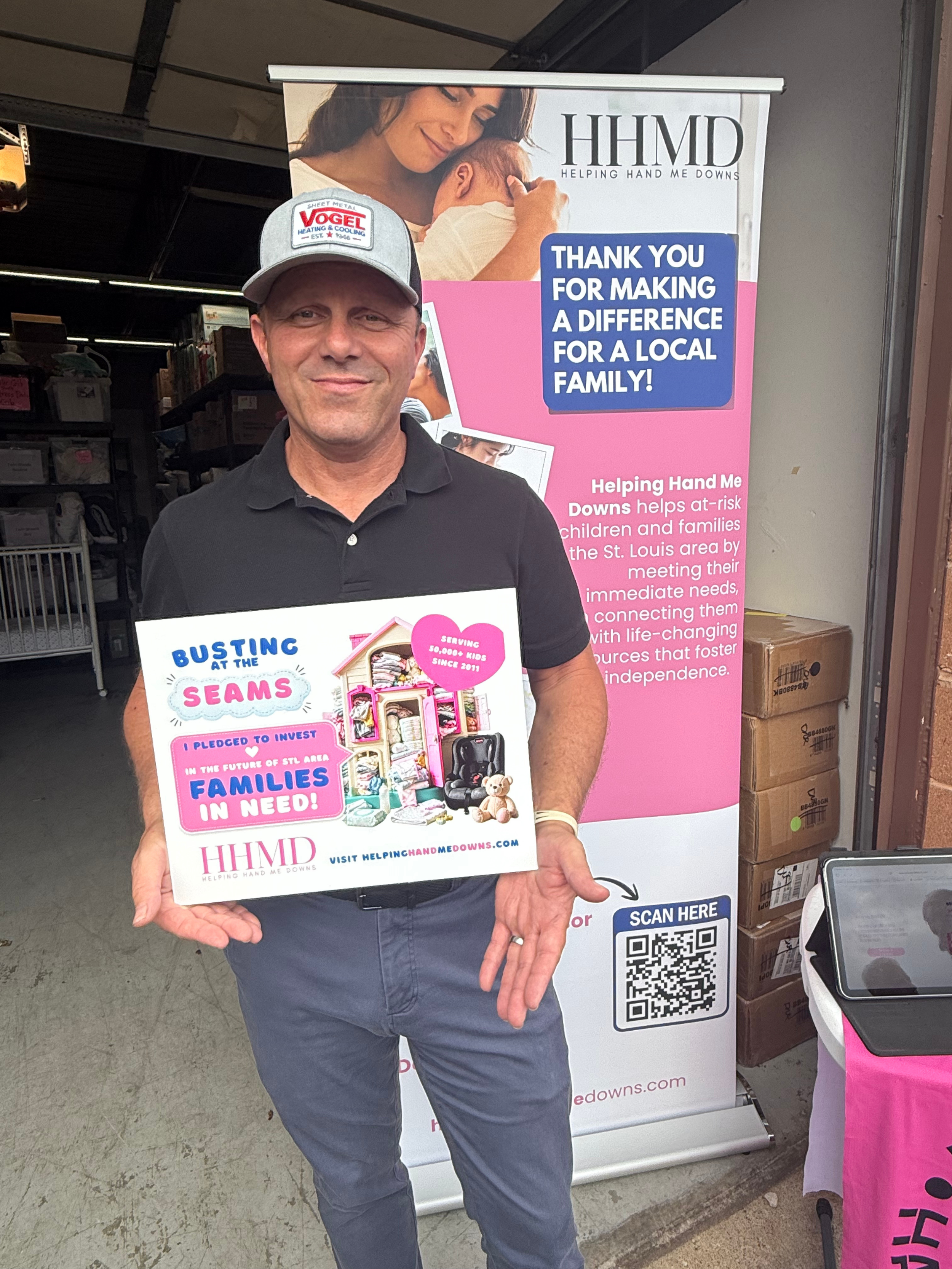 Man standing in front of a promotional banner for Helping Hand Me Downs, holding a sign promoting the organization. The banner features a message of thanks for supporting families with children in need. Helping Hand Me Downs Building Campaign.