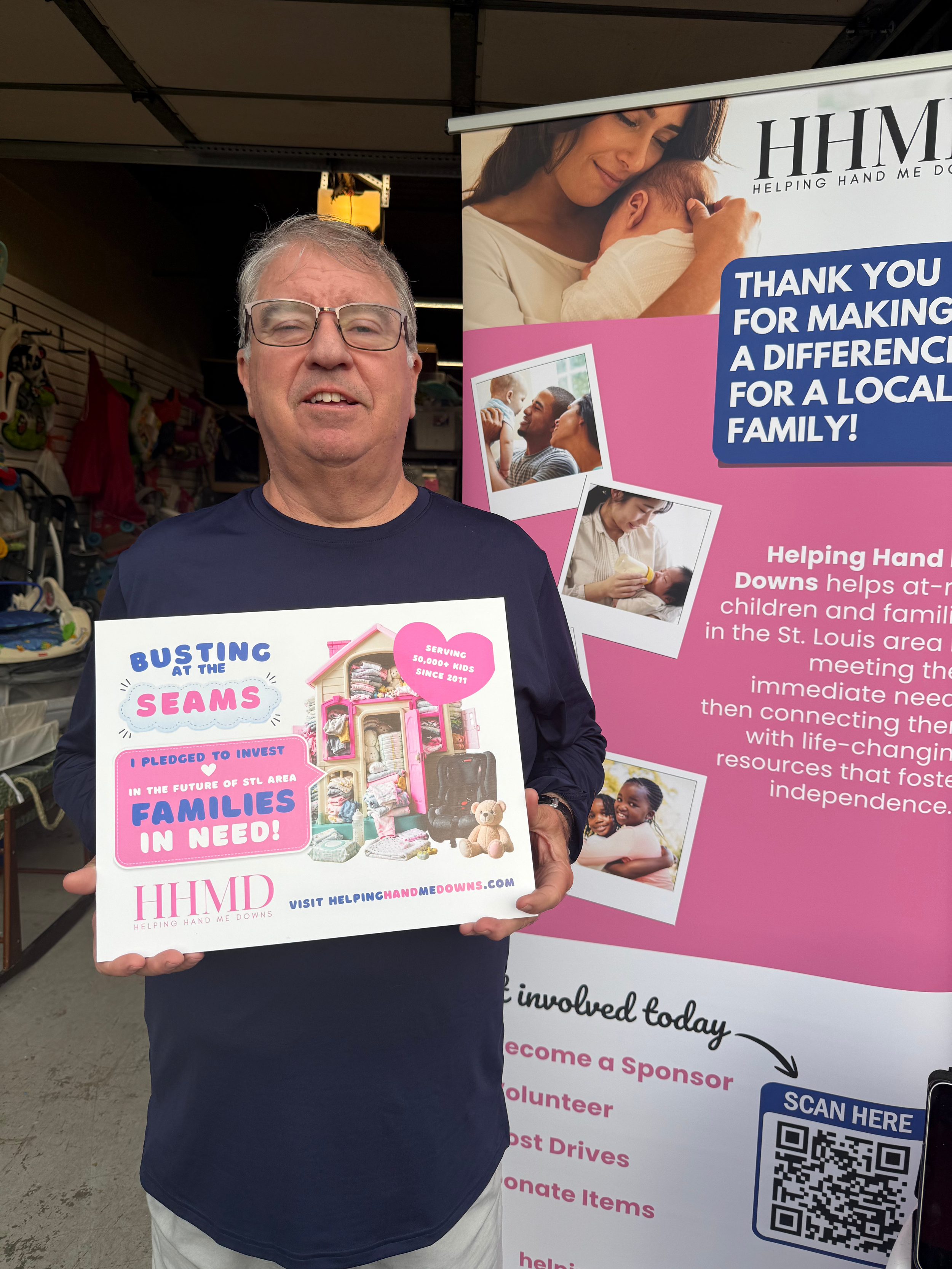 A man with glasses holding a sign promoting helping children in need, standing in front of a pink and white banner with images of families and children and text about community service. Helping Hand Me Downs Building Campaign Busting at the Seams.