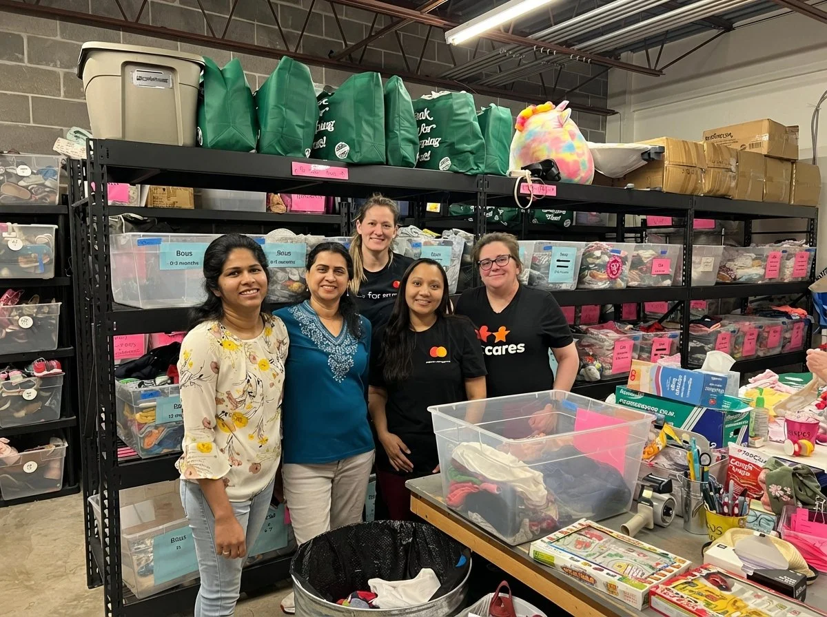 Five volunteers inside the Helping Hand Me Downs warehouse with shelves stocked with boxes, bins, and bags containing clothing and other supplies, sorting clothing and getting necessary items ready for St. Louis families in need.
