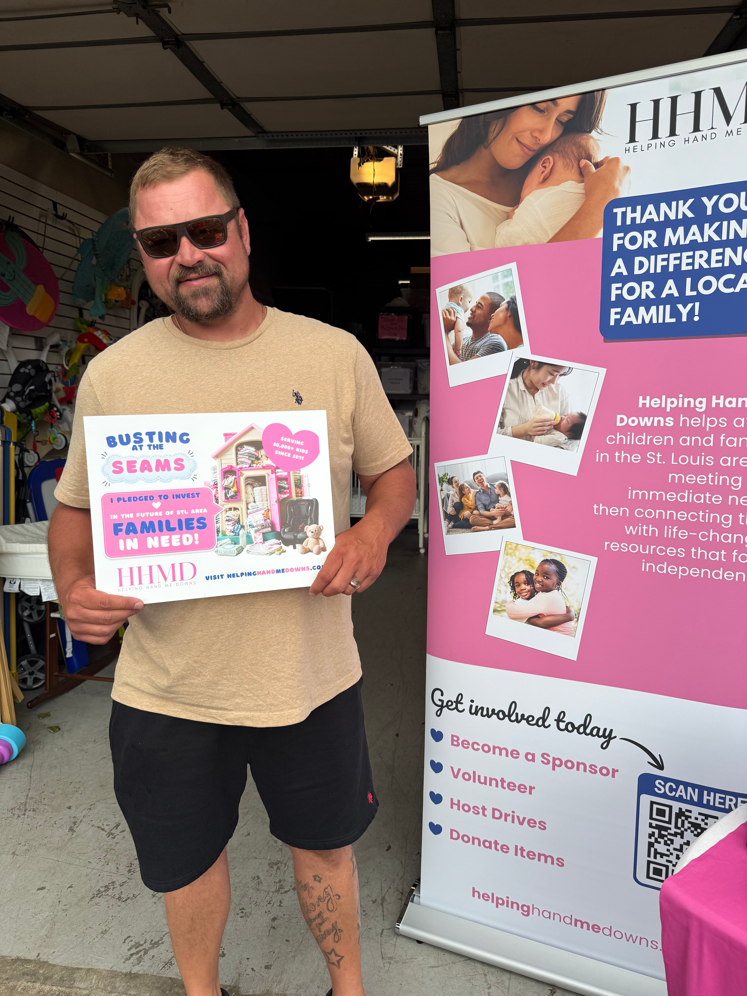 A smiling man wearing sunglasses and a tan t-shirt holding a flyer at an outdoor charity event, with a pink and white informational banner beside him. Helping Hand Me Downs Building Campaign Busting at the Seams.