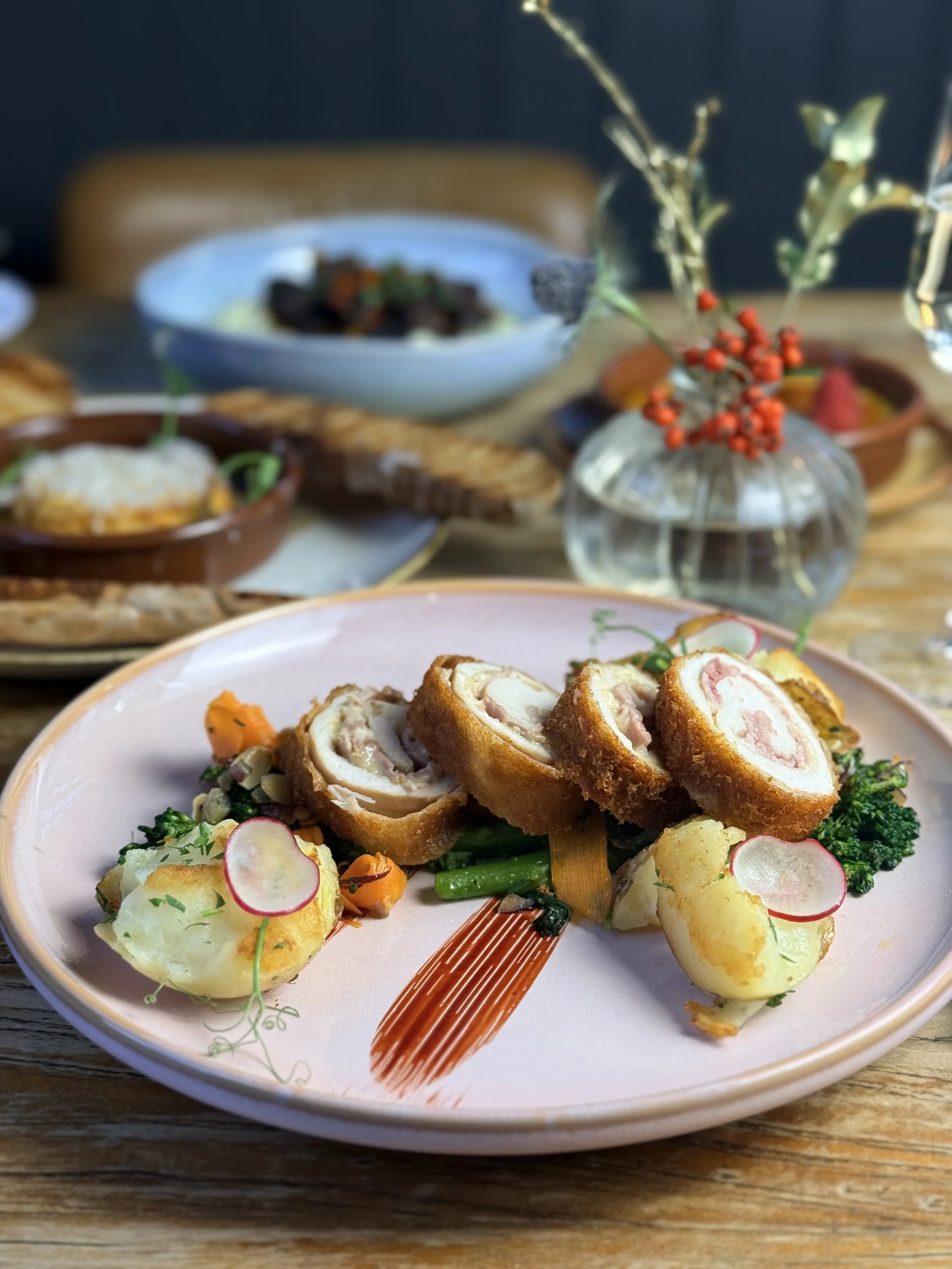 A plate of breaded and fried rolled meat with a pink filling, accompanied by mashed potatoes garnished with radish slices and vegetables, on a pink plate set on a wooden table with other dishes and a decorative glass vase in the background.