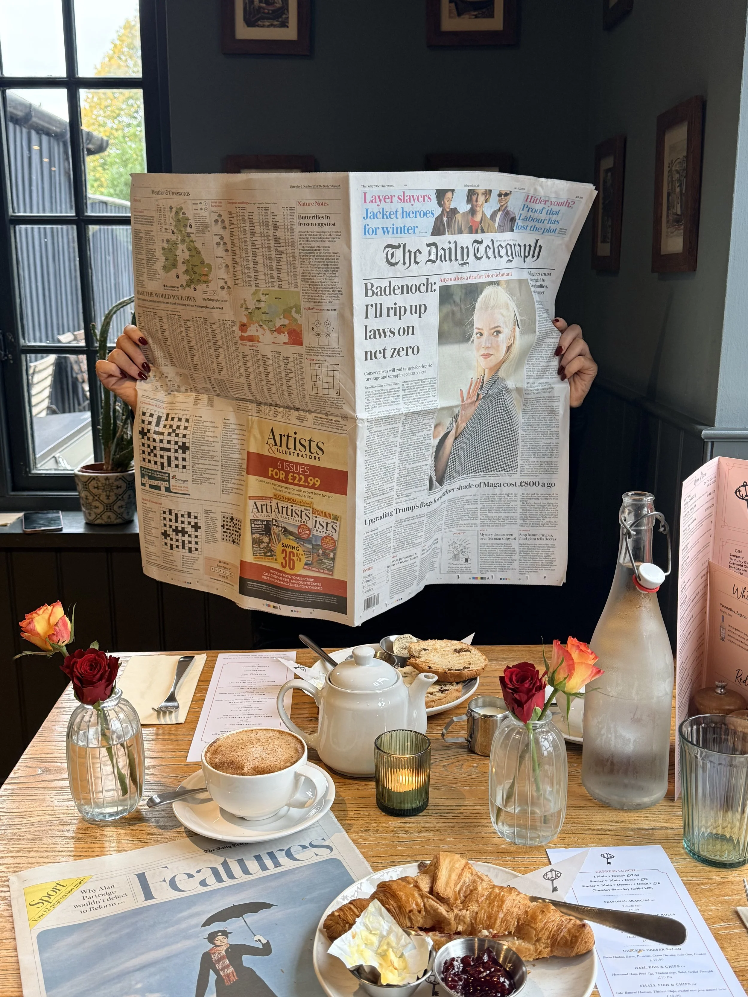 Person reading a newspaper at a breakfast table with coffee, croissants, flowers, and a water bottle.
