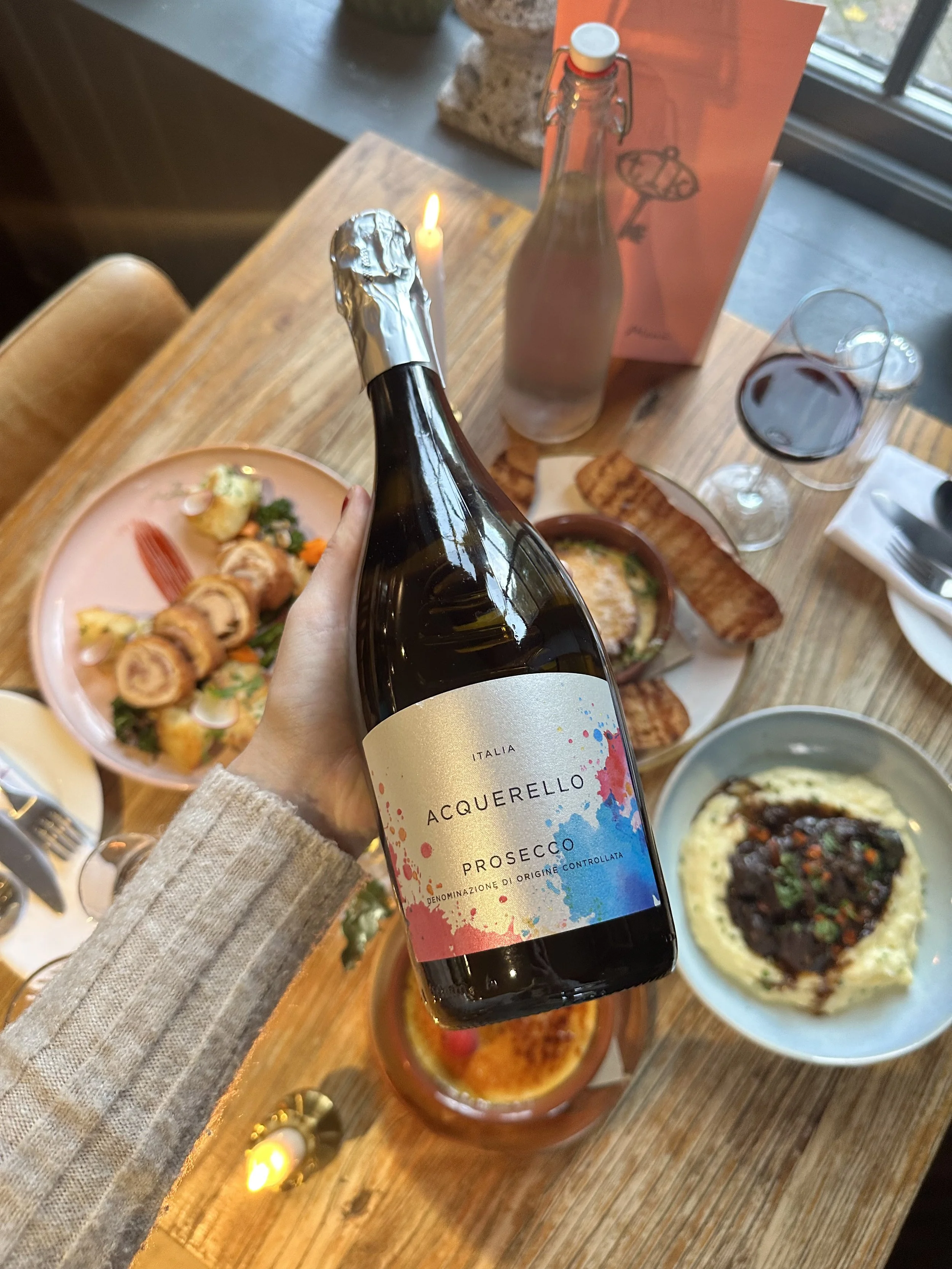 A hand holding a bottle of Acquerello Prosecco in front of a table with various dishes including a salad, a bowl of mashed potatoes or risotto topped with meat, and a plate with sliced meat and vegetables. There is also a glass of red wine, a bottle of water, and a pink paper bag on the table.