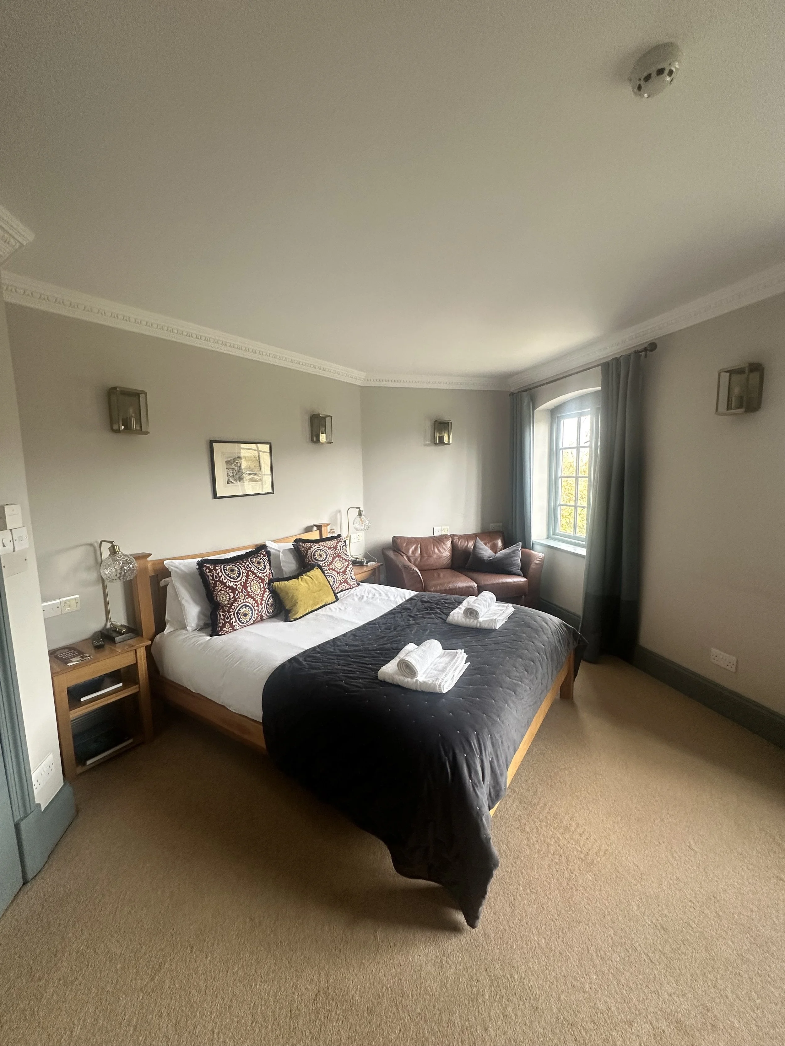 A cozy hotel room with a bed, decorative pillows, and neatly folded towels. There is a sofa by the window, and artwork on the wall. The room has beige carpet and soft lighting.