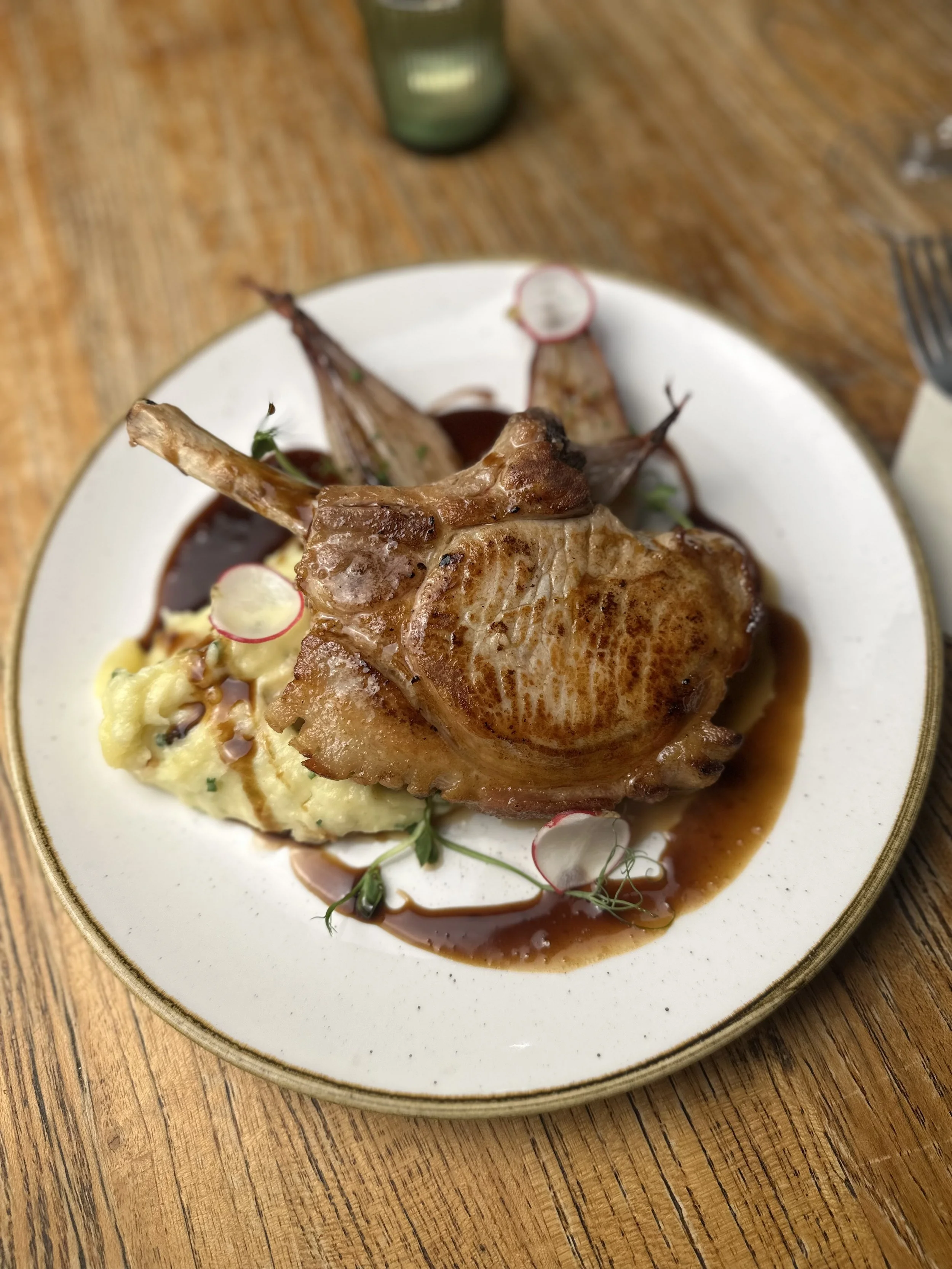 A plate of roasted lamb chops served with mashed potatoes, garnished with radish slices and microgreens, drizzled with sauce.