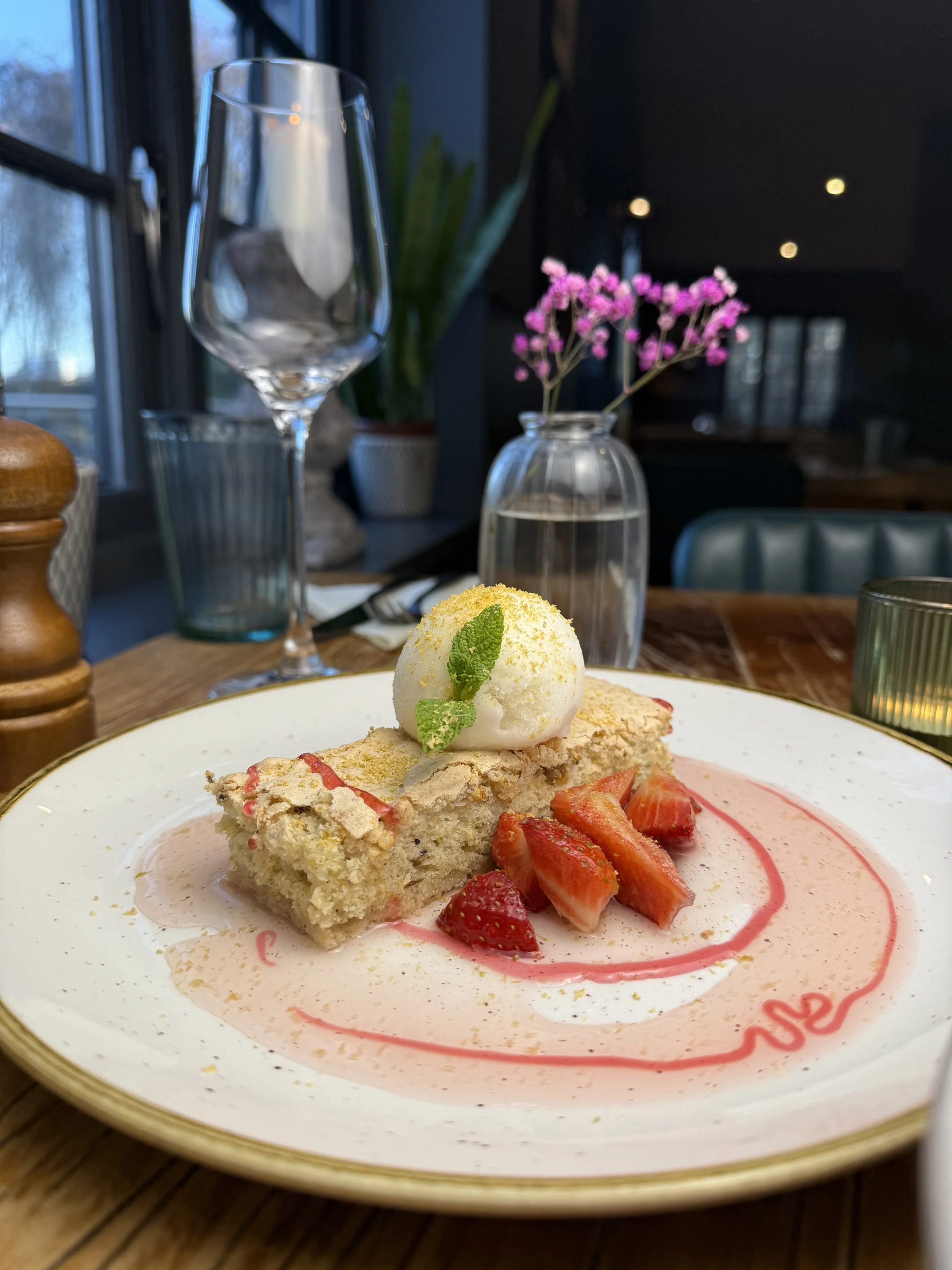 Slice of strawberry shortcake with ice cream, strawberries, and garnishes on a white plate with pink sauce, set on a wooden table in a cozy restaurant.