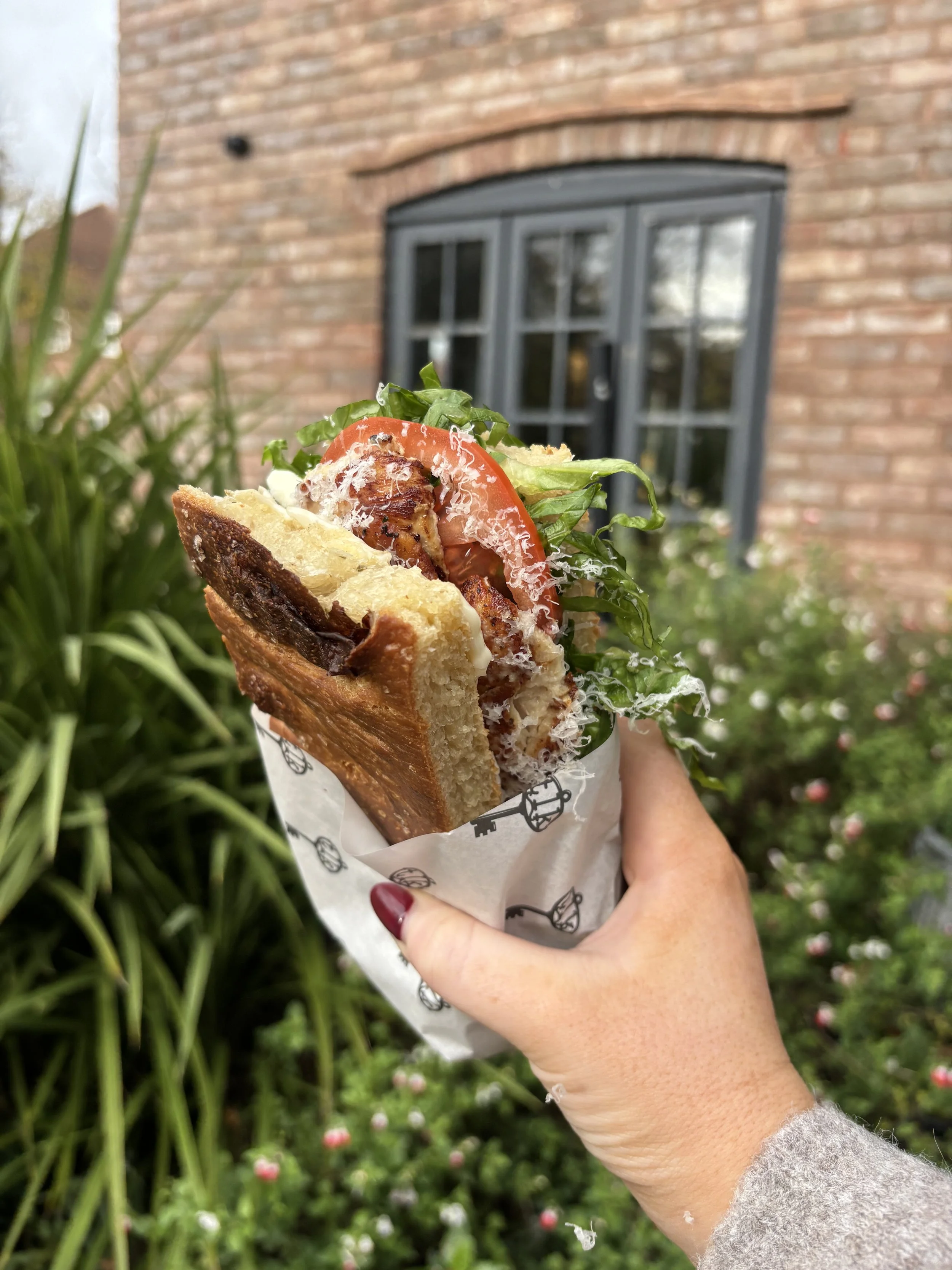 Hand holding a large sandwich with lettuce, tomato, grilled meat, cheese, and toasted bread outdoors with a brick house and window in the background.