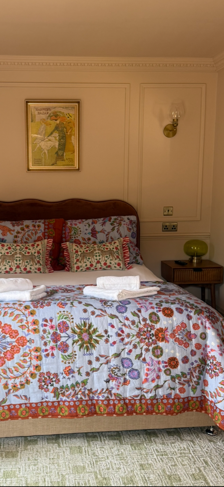 A neatly made bed with a floral bedspread, two decorative pillows with animals, and white towels on top. The bed is in a room with framed artwork, a wall sconce, and a nightstand with a green lamp.