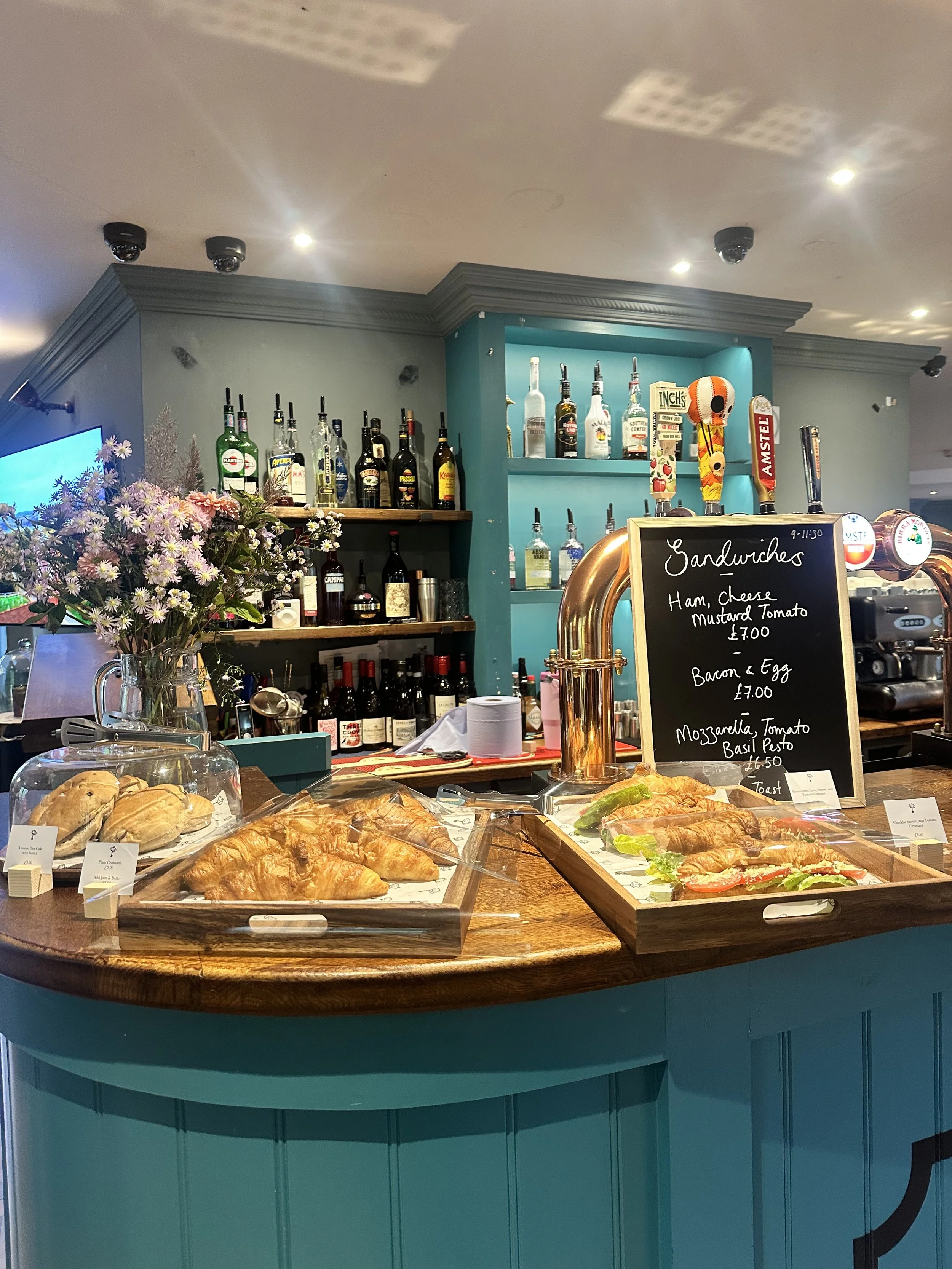 A counter with pastries and a chalkboard menu in a cafe or bar. The counter has croissants, sandwiches, and a vase of flowers. Behind are shelves with bottles of alcohol and a beer tap handle.