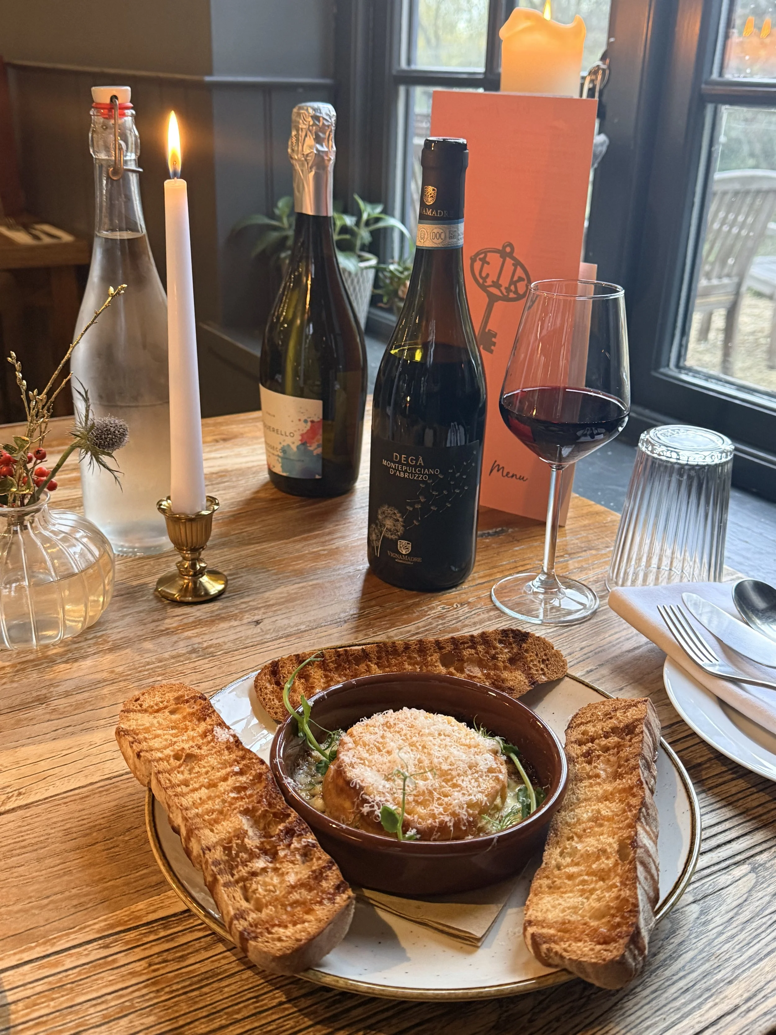 A table setting with a bowl of baked ricotta topped with cheese and herbs, surrounded by toasted bread slices, a glass of red wine, and bottles of sparkling wine. There is also a lit candle, a small floral arrangement, a menu, and a glass water bottle on the wooden table inside a cozy restaurant.