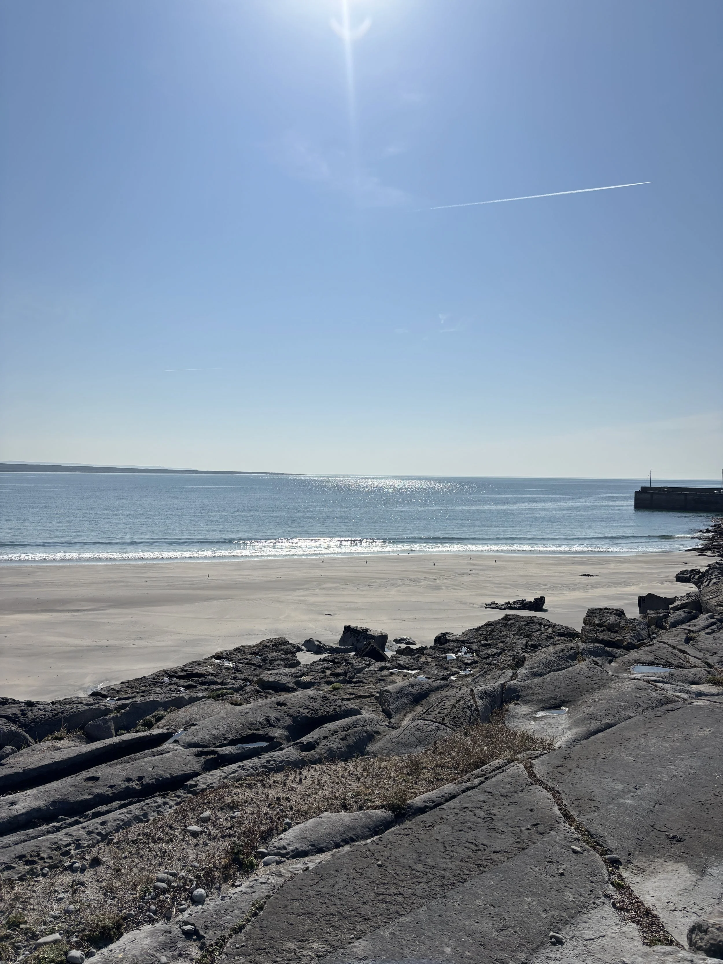 View of a sandy beach with large rocks in the foreground, calm ocean water, clear blue sky, and a distant landmass along the horizon. Bright sun overhead with a slight contrail in the sky.
