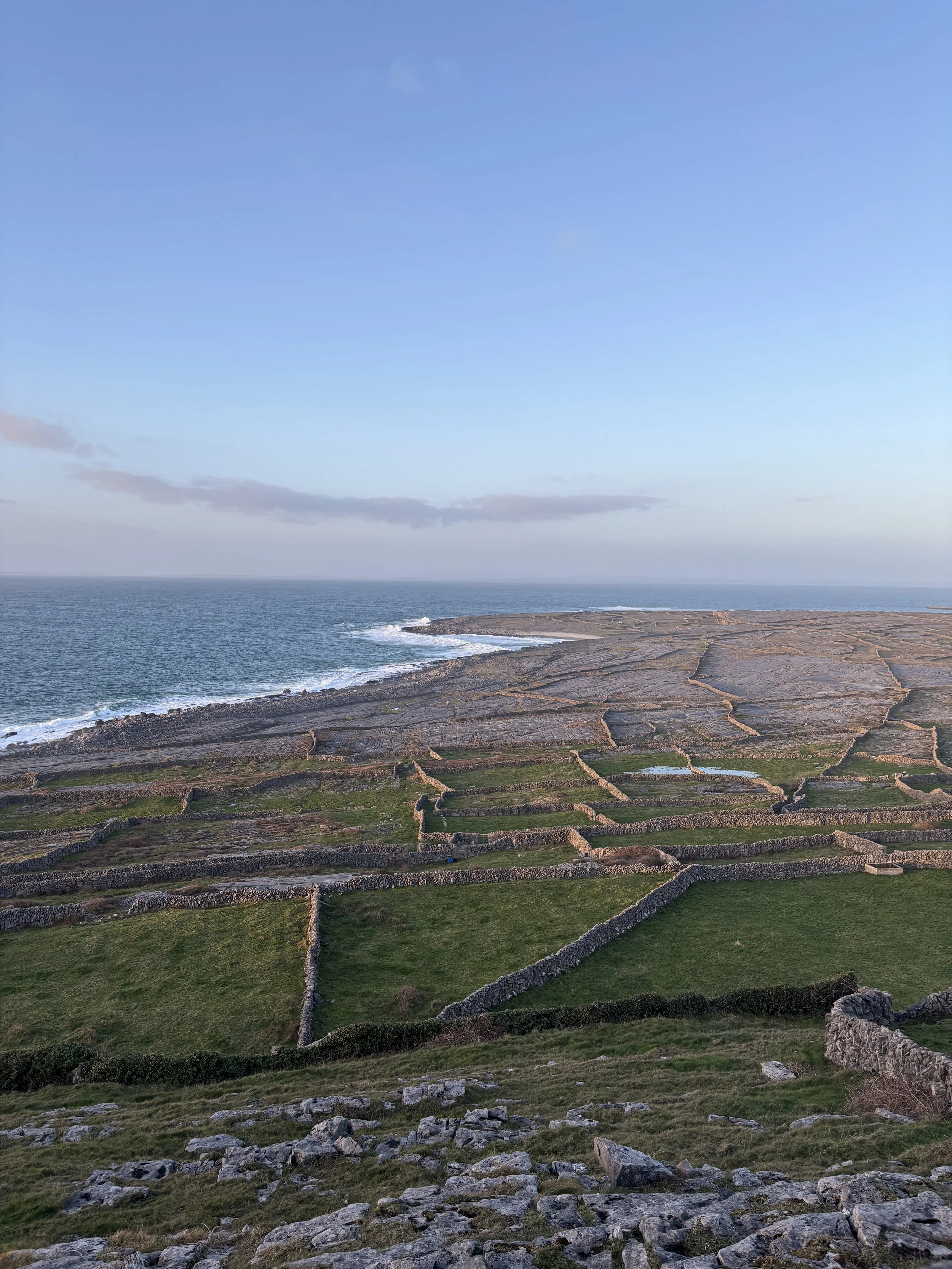 A rocky shoreline with terraced stone walls and patches of green grass, leading to the ocean under a mostly clear blue sky with some thin clouds.