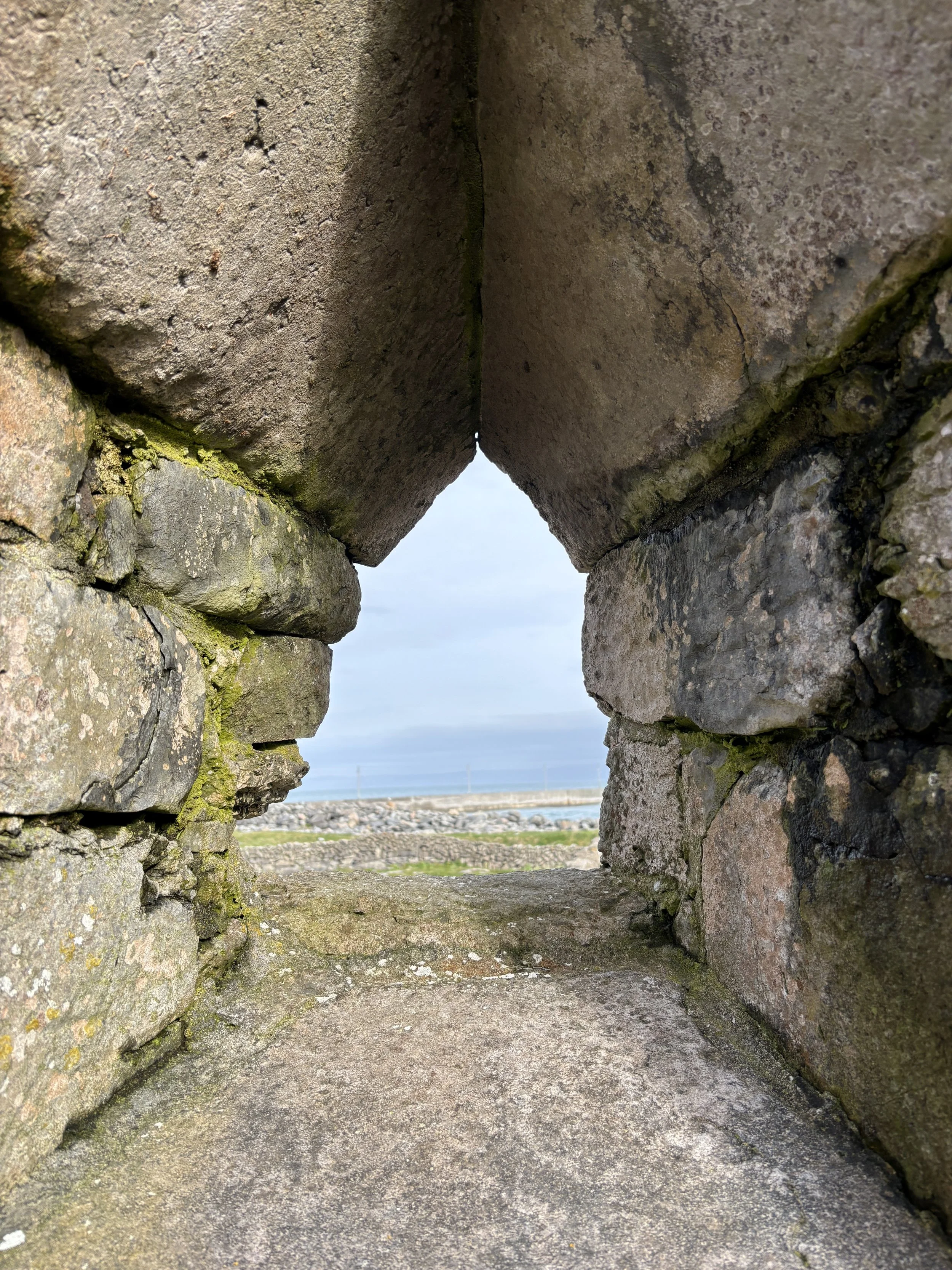 View through a small opening in a stone wall, with a distant view of the sky and landscape beyond.