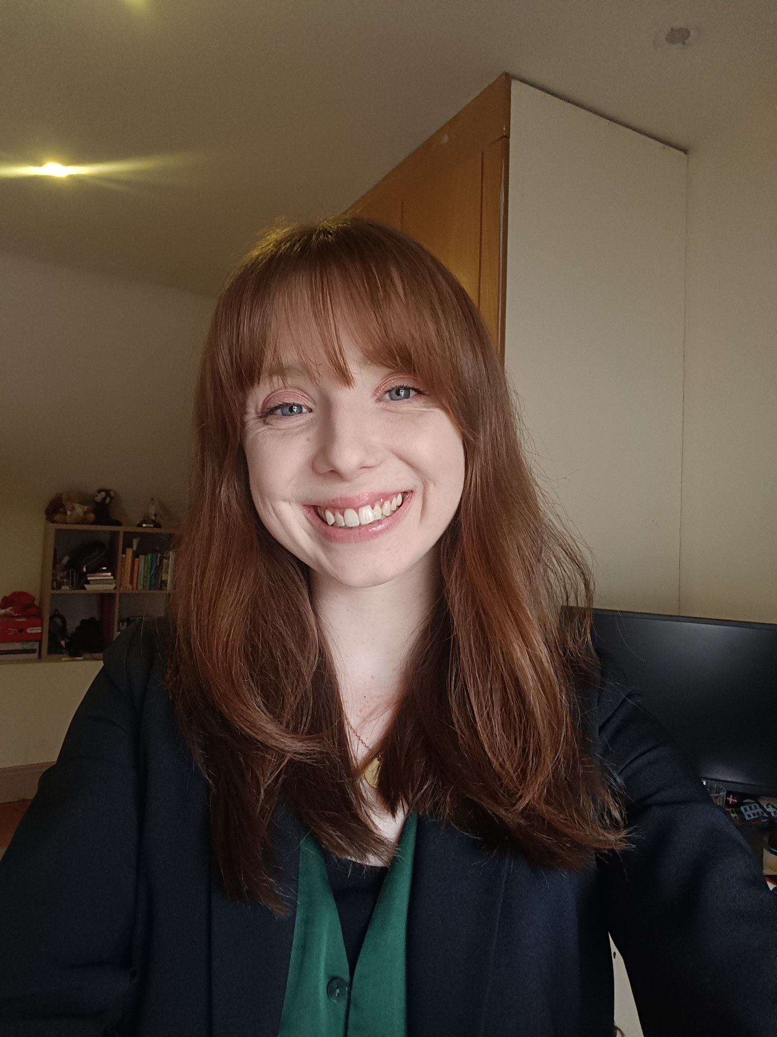 A woman with long red hair smiling, wearing a dark blazer and green blouse, sitting in a room with bookshelves and a television in the background.