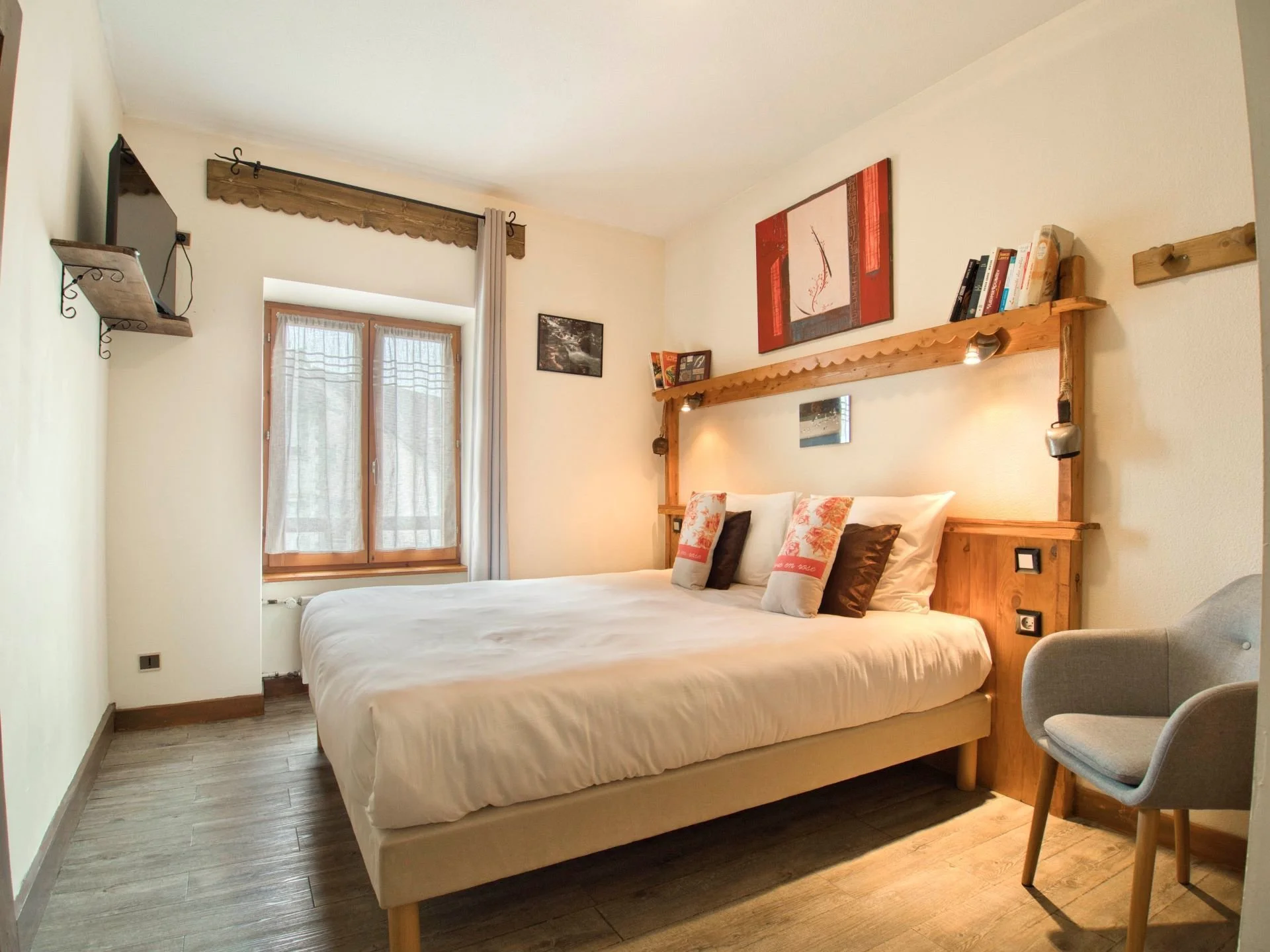 Chambre avec lit double, fenêtres, décor en bois, tableaux, étagères avec livres et lampes, fauteuil gris à côté du lit.