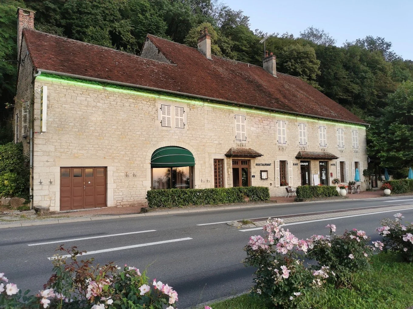 Bâtiment en pierre avec plusieurs fenêtres à volets blanc, un restaurant avec une terrasse extérieure, des fleurs en avant-plan, et un feuillage dense derrière, situé au bord de la route.