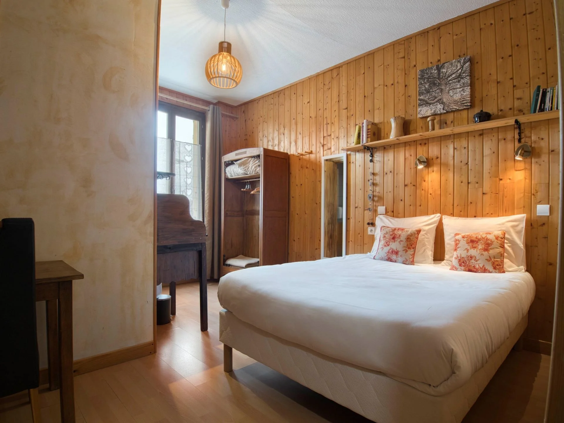 Chambre à coucher avec lit double, murs en bois, décor simple, fenêtres avec rideaux, étagère avec livres et décorations, lampe suspendue, tableau d'arbre sur le mur, éclairage doux.