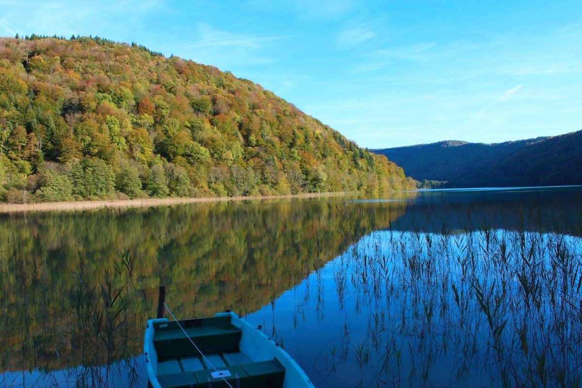 Un lac entouré de collines boisées avec un bateau blanc à l'avant, reflet des arbres dans l'eau, et un ciel bleu clair.