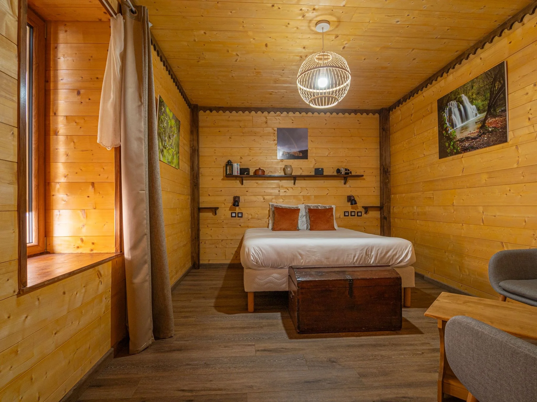 Chambre en bois avec lit, tableaux de nature, étagères, fenêtre, fauteuils et coffre en bois.