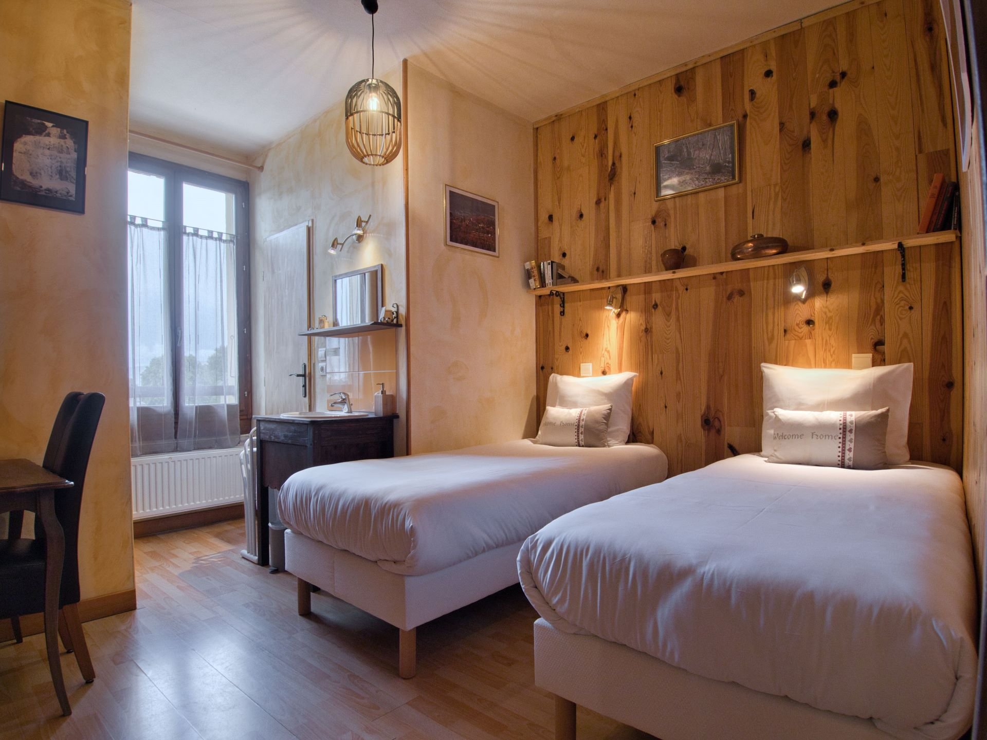 Chambre avec deux lits simples en bois clair, mur en bois derrière les lits, tableaux sur le mur, petite étagère avec décorations, fenêtre avec rideaux, petit lavabo avec miroir, chaise noire à gauche