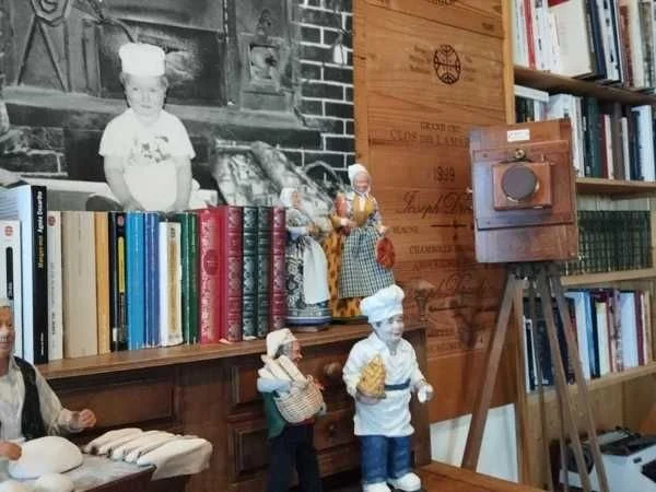 Exposition avec des figurines en bois habillées en costumes traditionnels et une photo en noir et blanc d'une petite fille en chef de cuisine. Étagères avec des livres et une vieille radio sur un trépied.