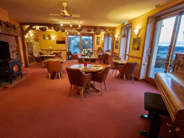 Intérieur d'un salon ou restaurant avec tables rondes, chaises en velours rose, cheminée à bois, piano en bois près d'une grande fenêtre, décorations murales, vue vers l'extérieur avec paysage