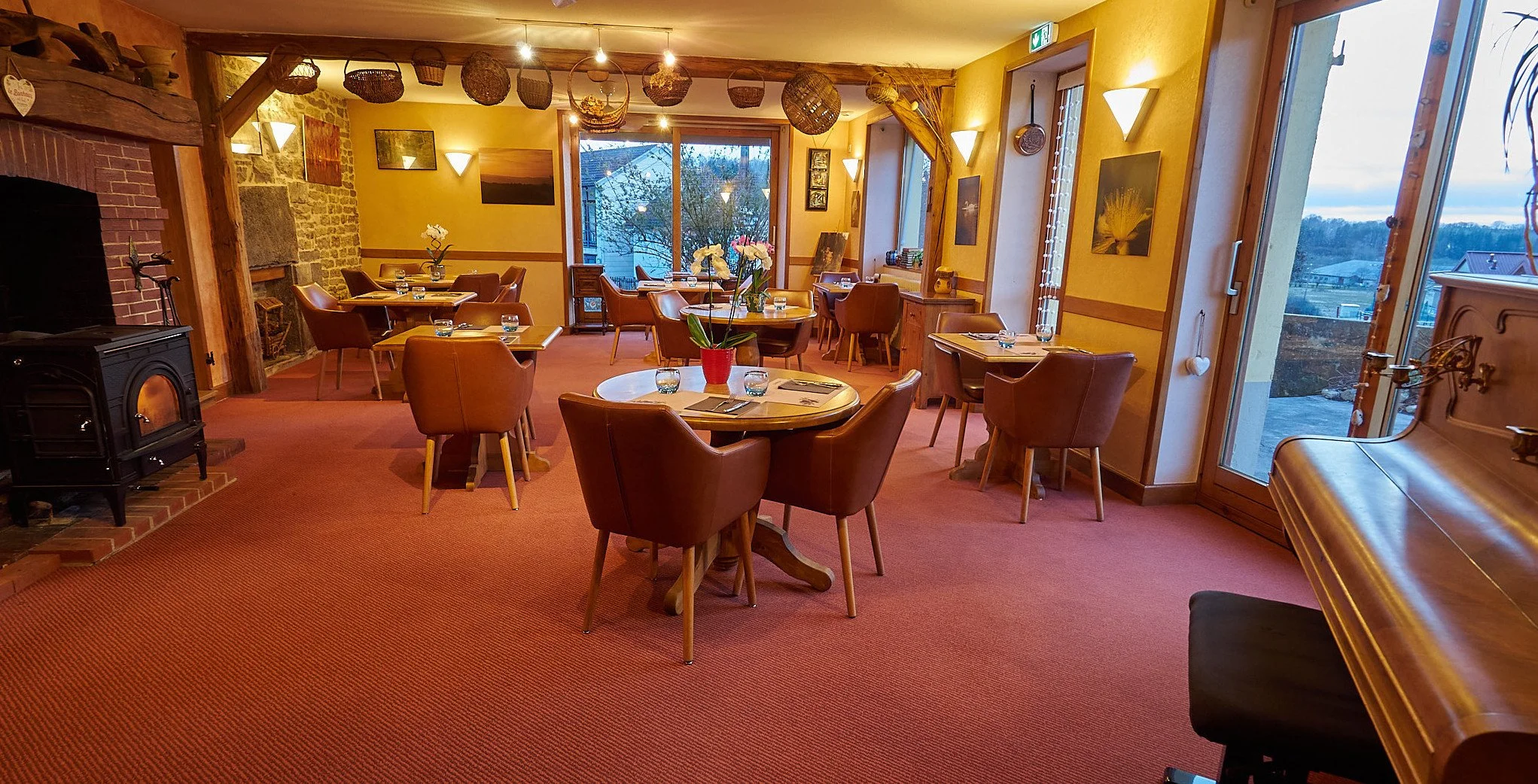 Une salle à manger chaleureuse avec plusieurs tables en bois, chaises en cuir marron, un poêle à bois noir, des décorations en paniers suspendus, des murs jaune avec des tableaux, des fenêtres laissant entrer la lumière naturelle, et un piano en bois.