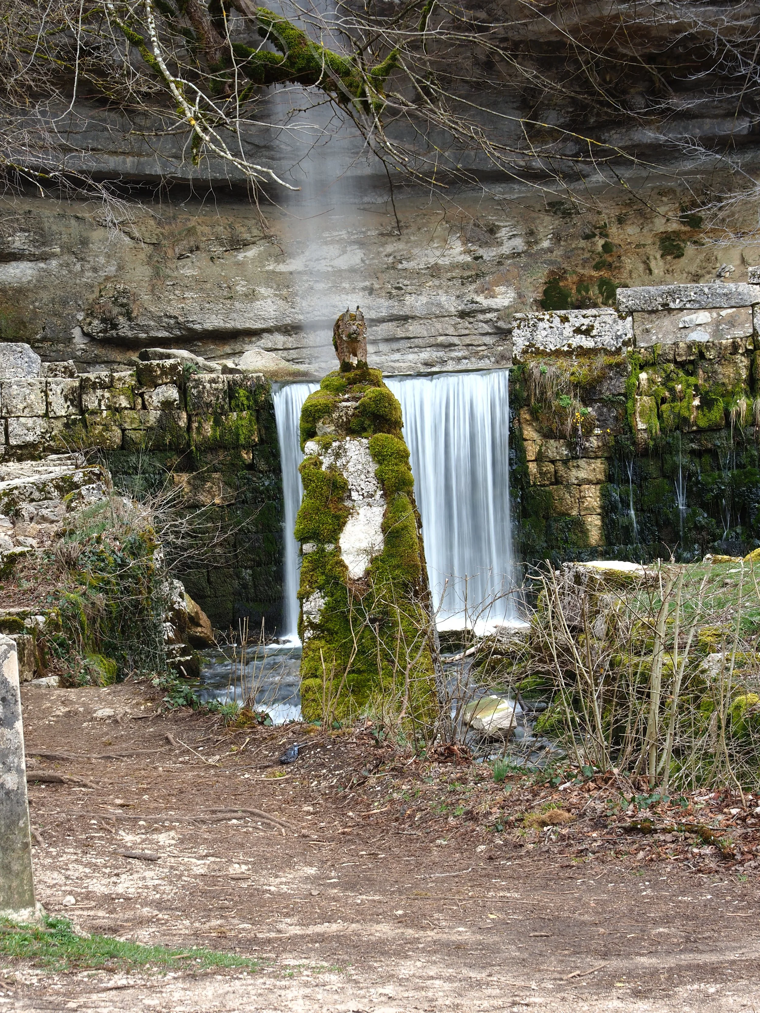 Cascade d'eau avec une structure recouverte de mousse dans un environnement naturel, entourée de roches, de branches et de végétation.