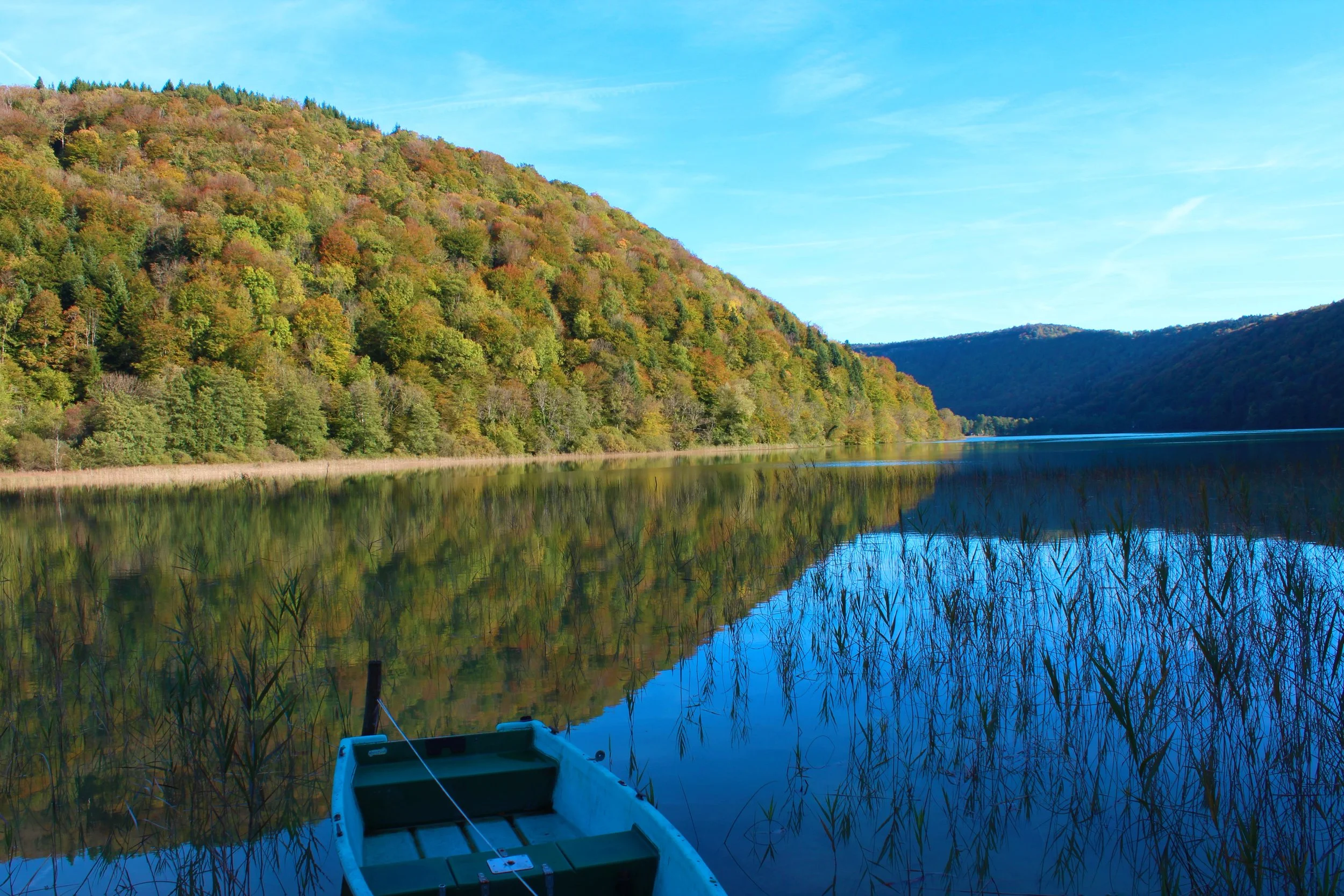 Une barque sur un lac entouré de collines boisées, avec des arbres dont les feuilles commencent à changer de couleur, reflet dans l'eau claire sous un ciel bleu.