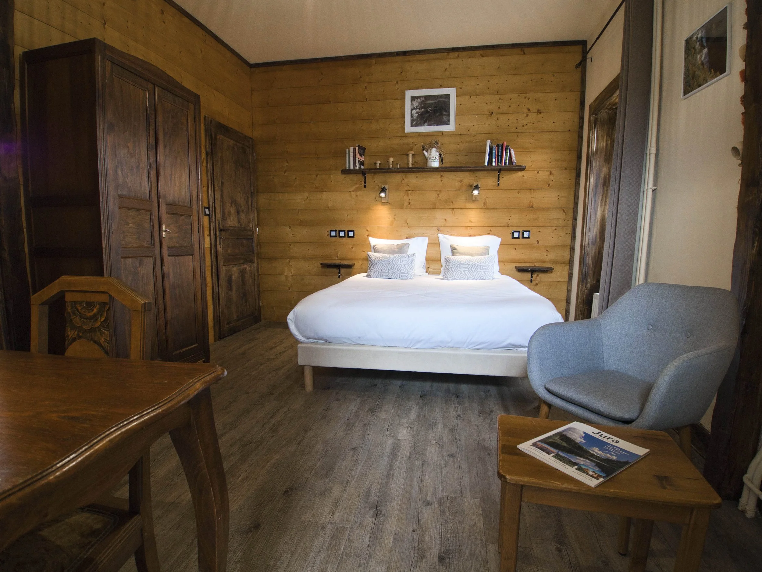 Chambre avec lit double, armoires en bois, fauteuil gris, table en bois avec magazine, décoration en bois et photos sur le mur.