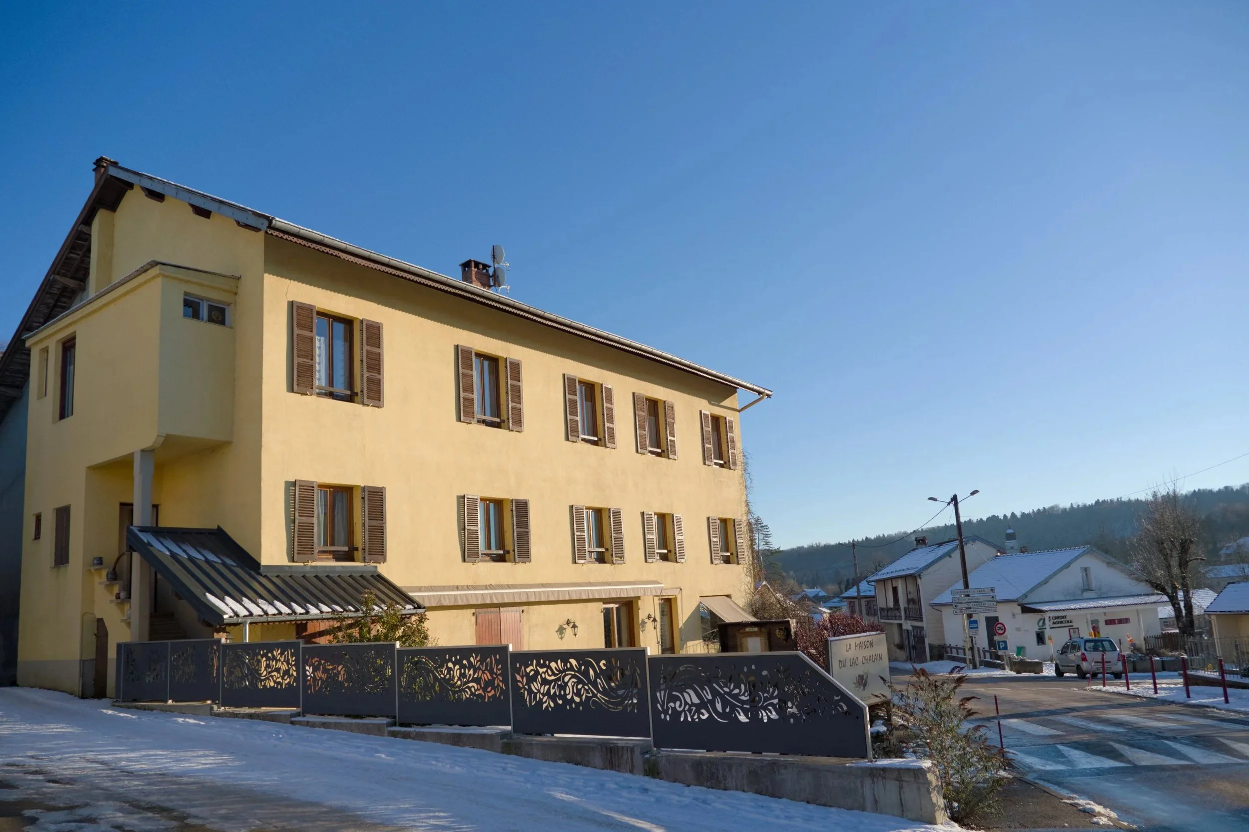 bâtiment résidentiel jaune avec plusieurs fenêtres à volets en bois, sous un ciel bleu clair, avec une rue enneigée devant.