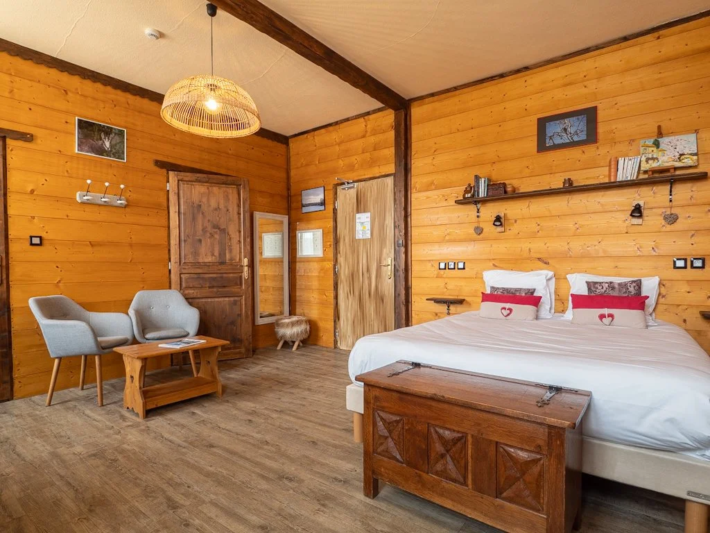 Une chambre avec murs en bois, un lit double avec des oreillers décoratifs, une étagère avec des livres, deux fauteuils en tissu gris, une table en bois, et une porte en bois. Décorations murales et un éclairage suspendu en rotin.