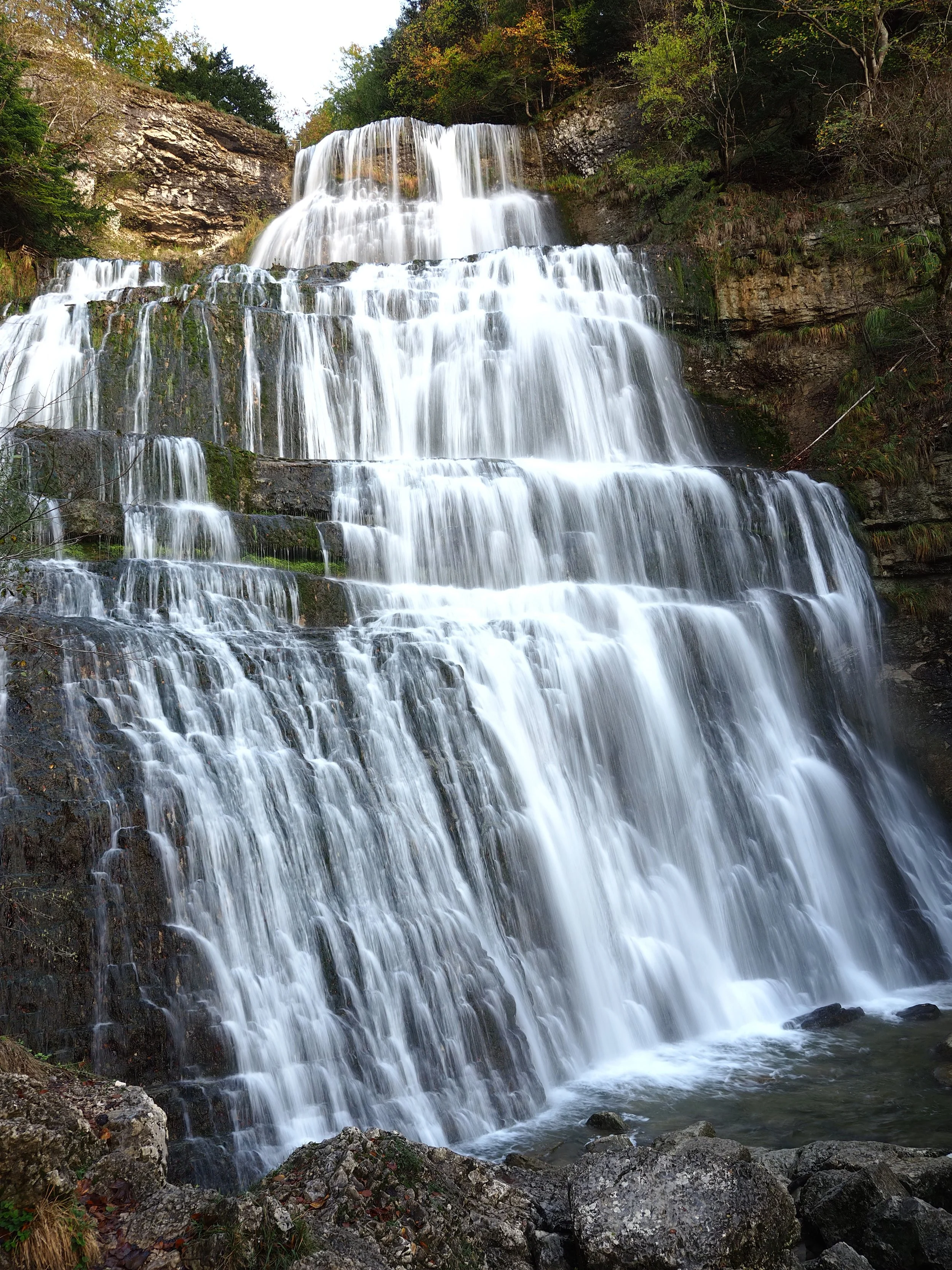 Une cascade d'eau tombant en plusieurs niveaux dans un environnement naturel avec des arbres verts et des rochers au premier plan.