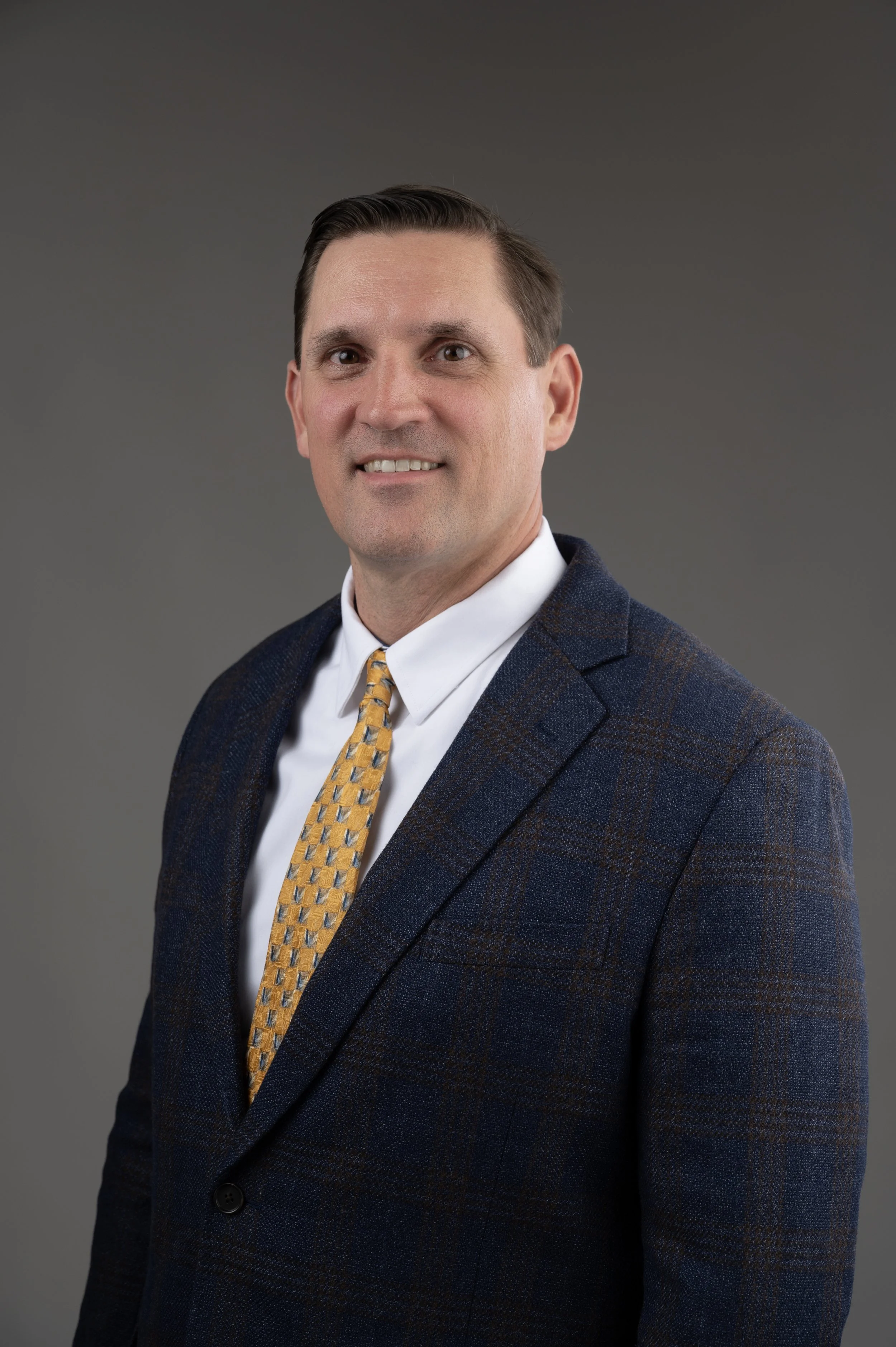 Professional headshot of a man in a suit with a navy blazer, white shirt, and plaid tie, smiling against a plain gray background.