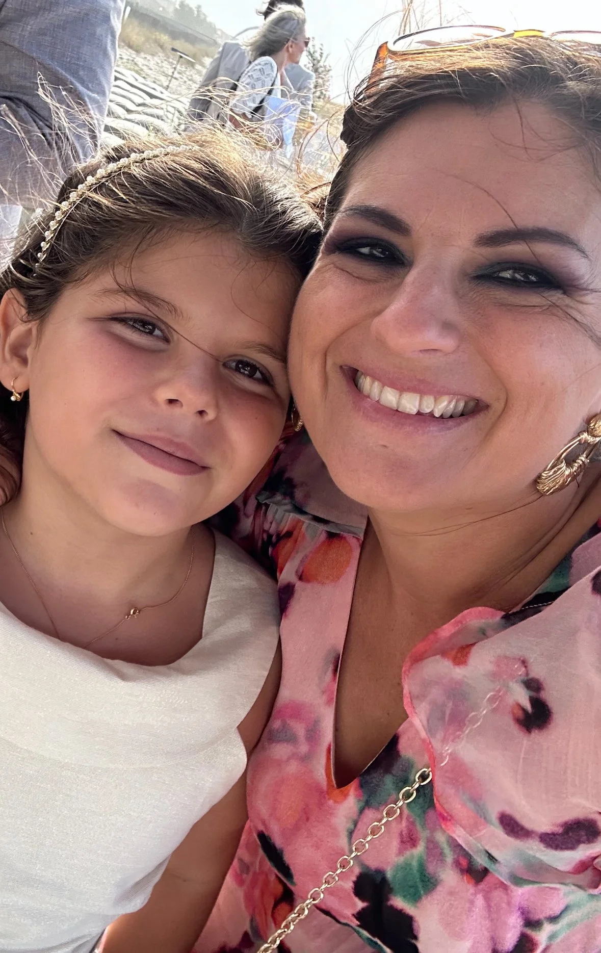 Close-up of two women smiling, leaning their heads together outdoors with a rocky terrain and a person in the background.