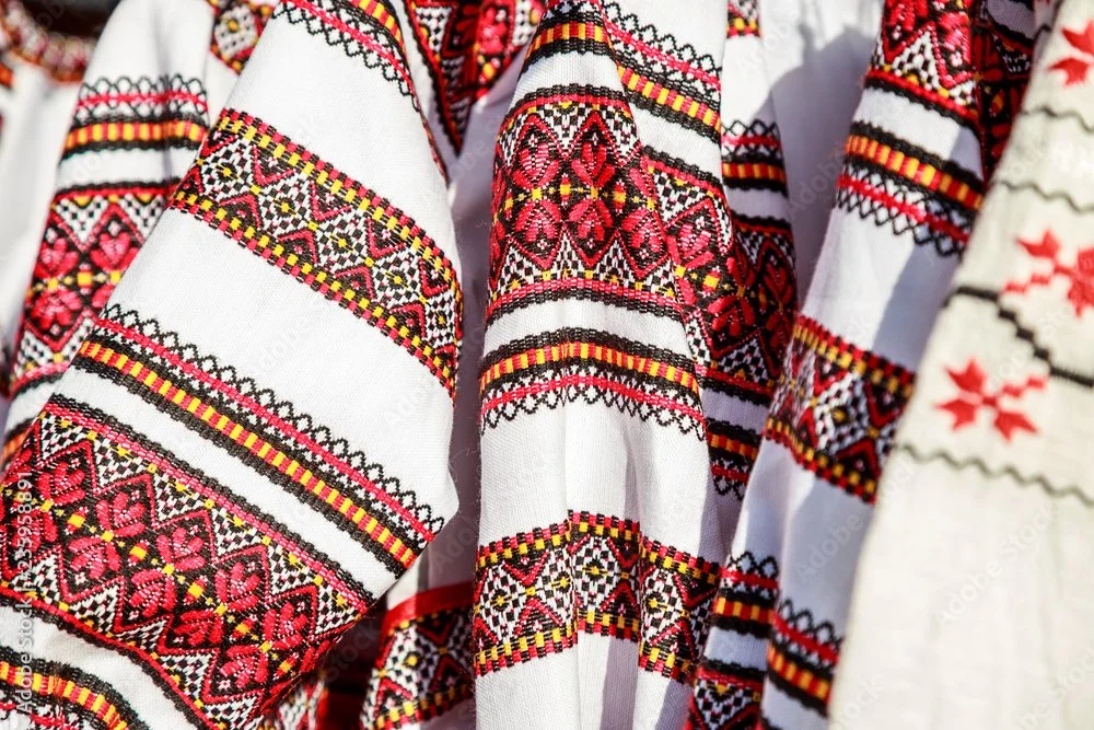 Close-up view of traditional embroidered fabric with intricate red, black, yellow, and white patterns, likely part of a traditional garment or textile.